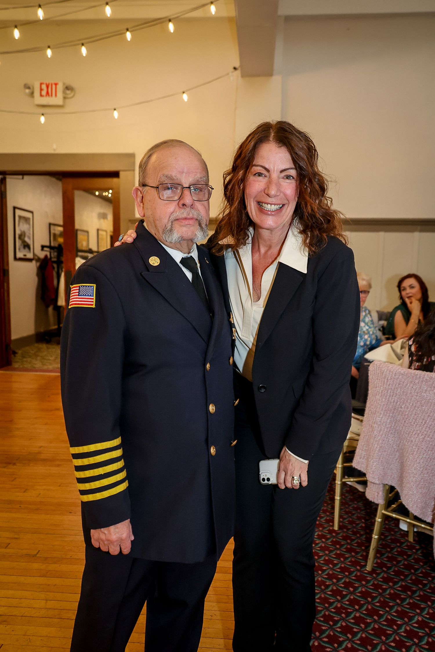 A fire chief in dress uniform poses with a smiling woman in a black blazer at a formal indoor event.
