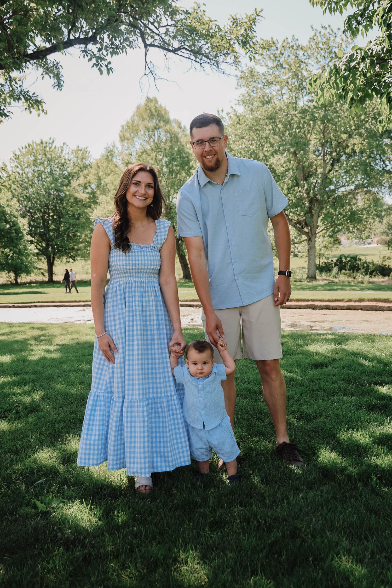 A young family with a baby stands on green grass in a sunny park, wearing coordinating blue outfits.