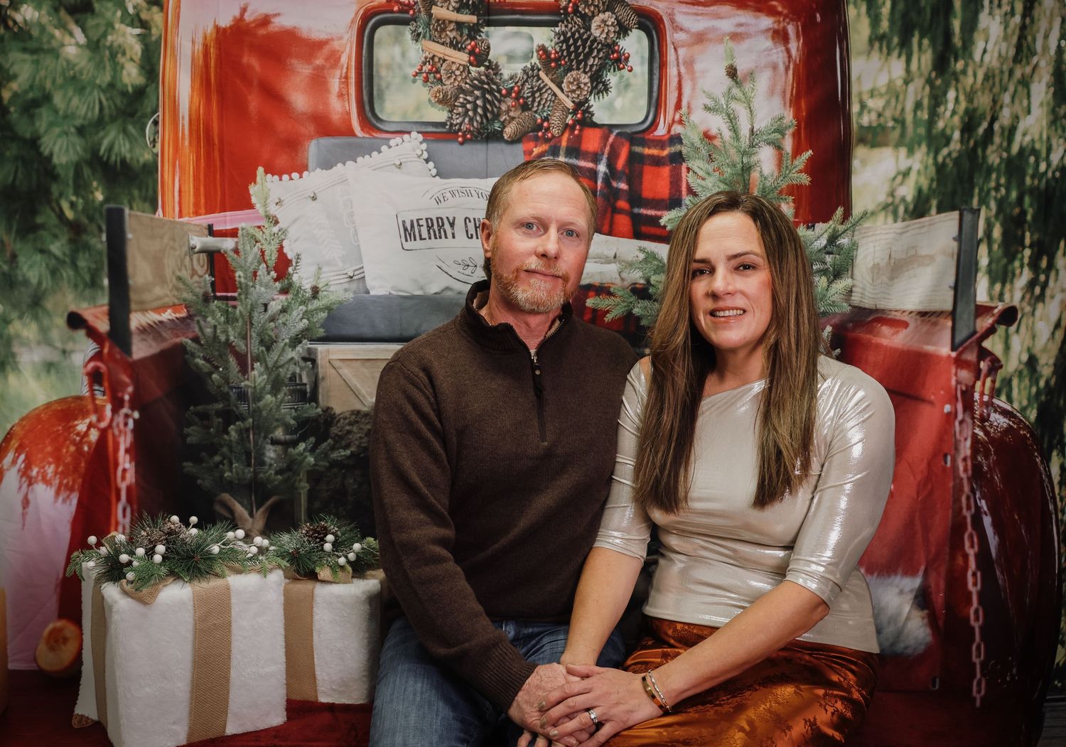 A couple smiles together in front of a festive Christmas backdrop with a red truck and holiday decorations.