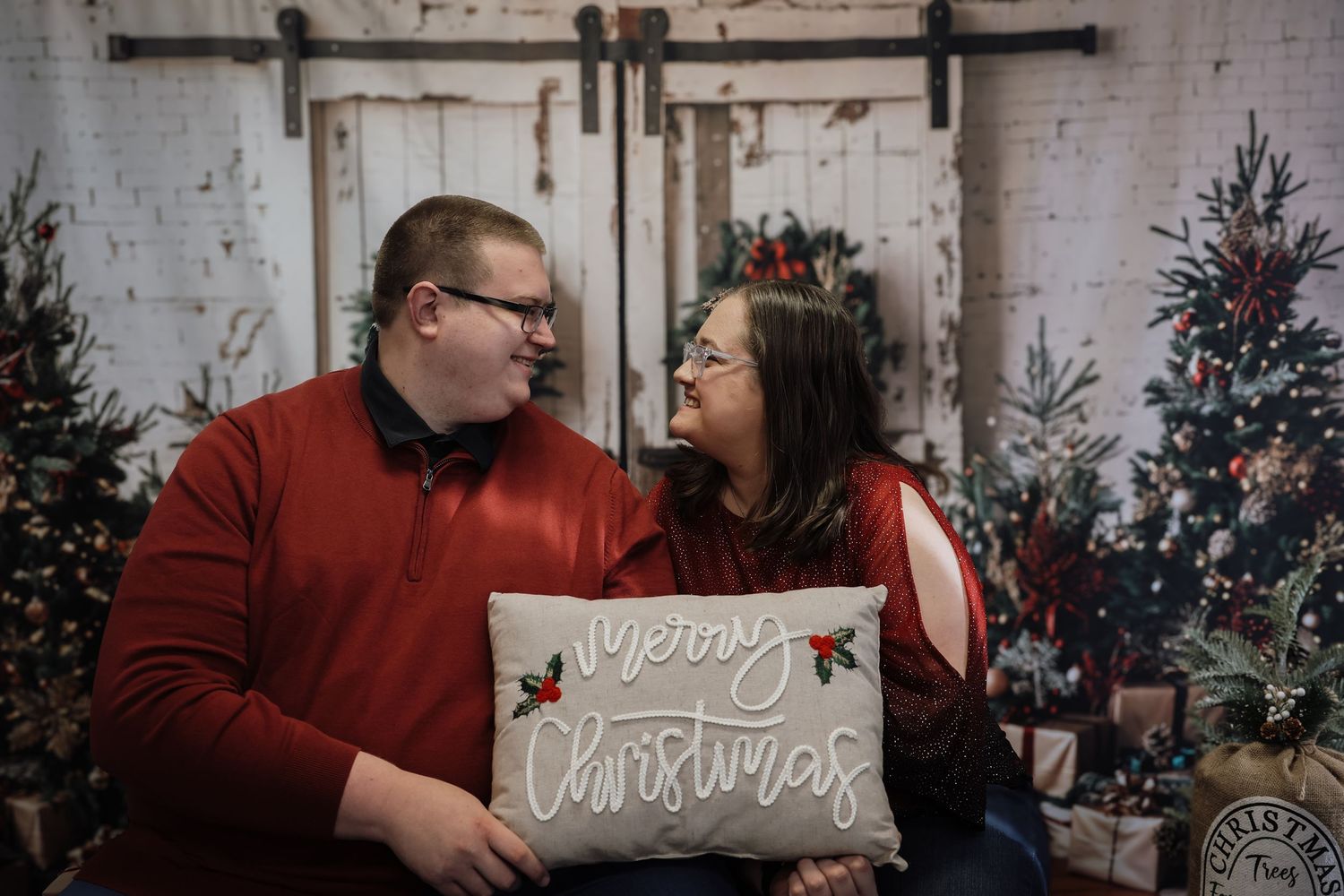 Couple holding a Merry Christmas pillow smiling at each other in front of festive holiday trees and barn door backdrop.