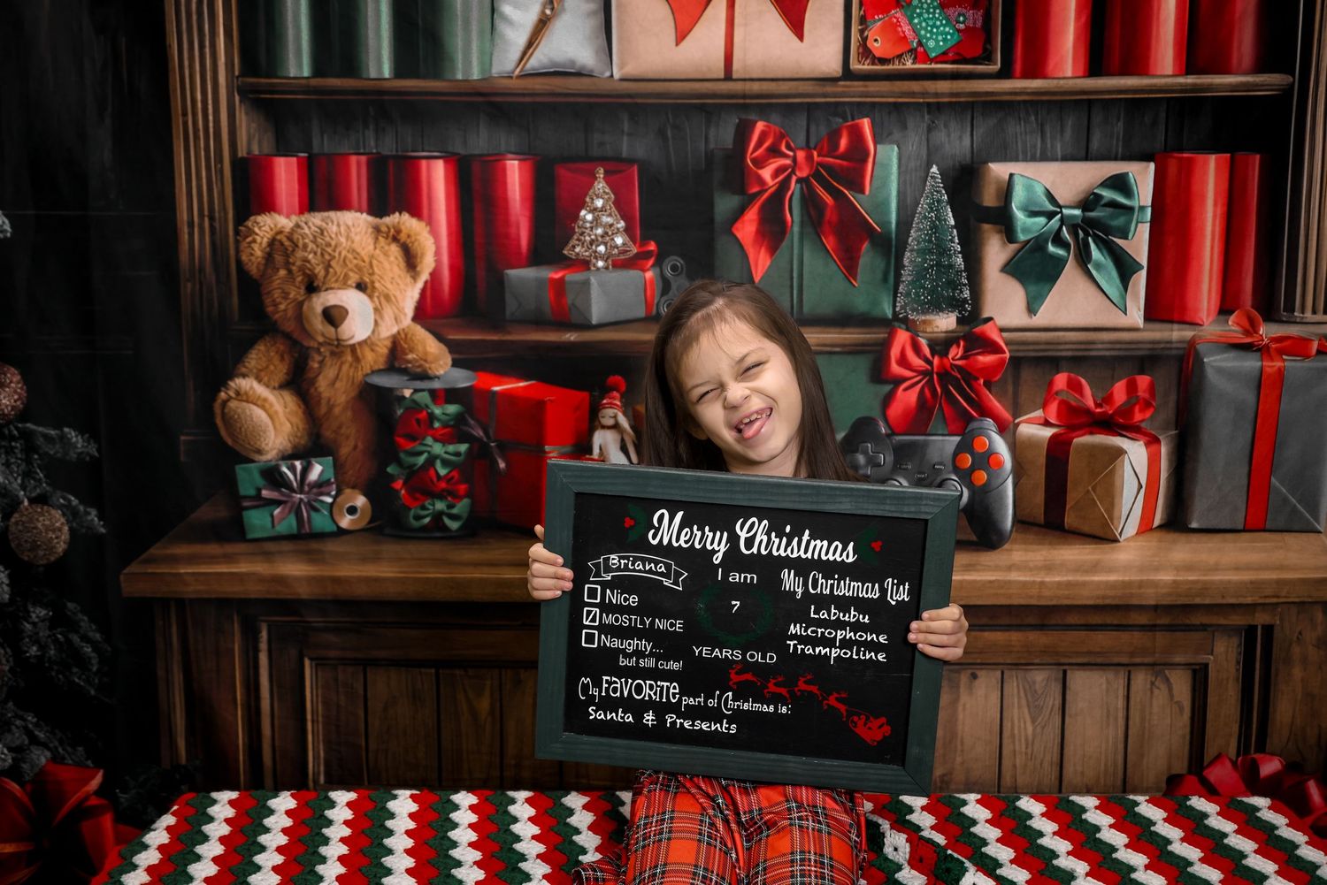 Young girl holding a Merry Christmas chalkboard sign surrounded by festive gifts, teddy bear, and holiday decorations.
