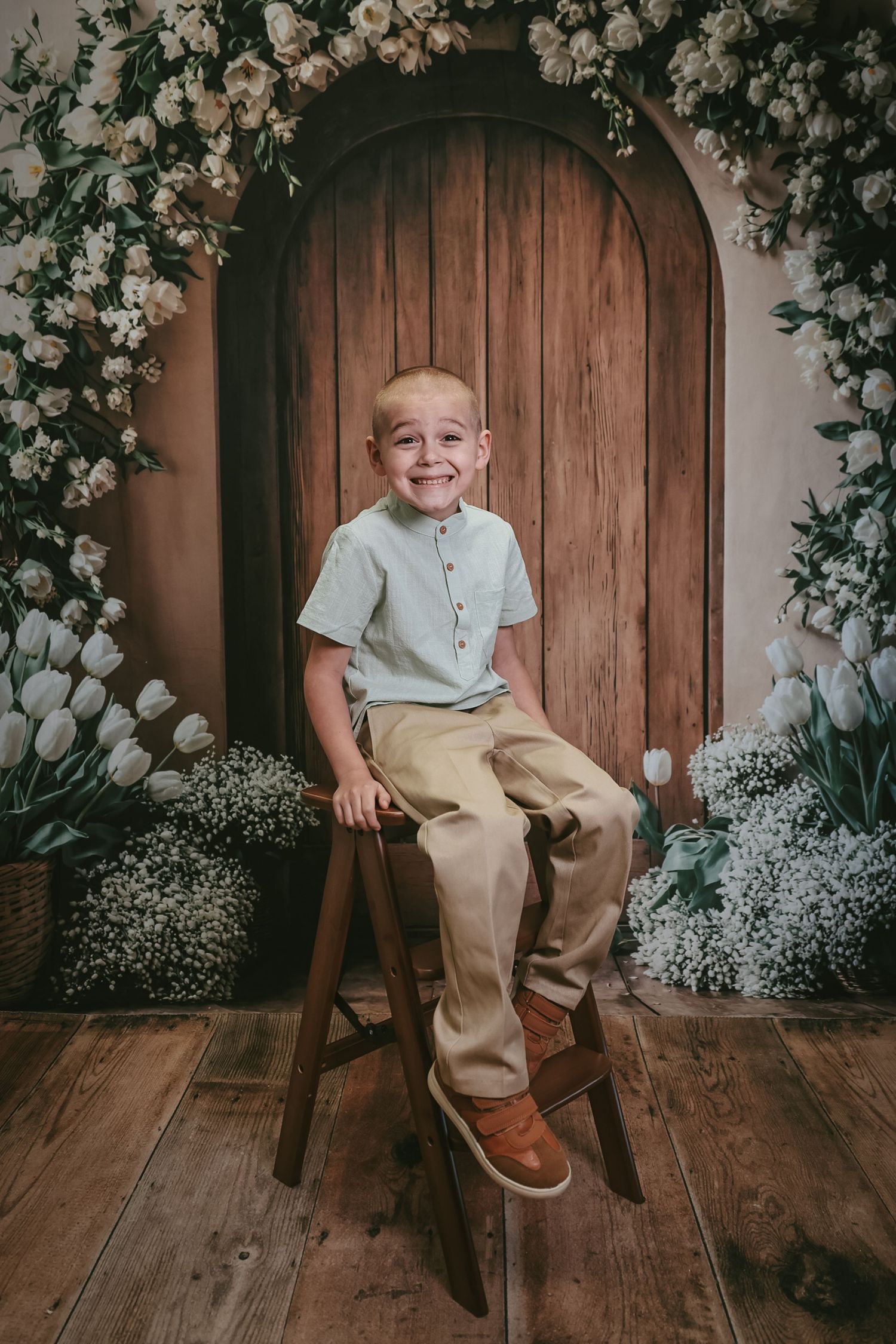 Smiling boy in light blue shirt and khaki pants sits on wooden stool before floral arch with white tulips.
