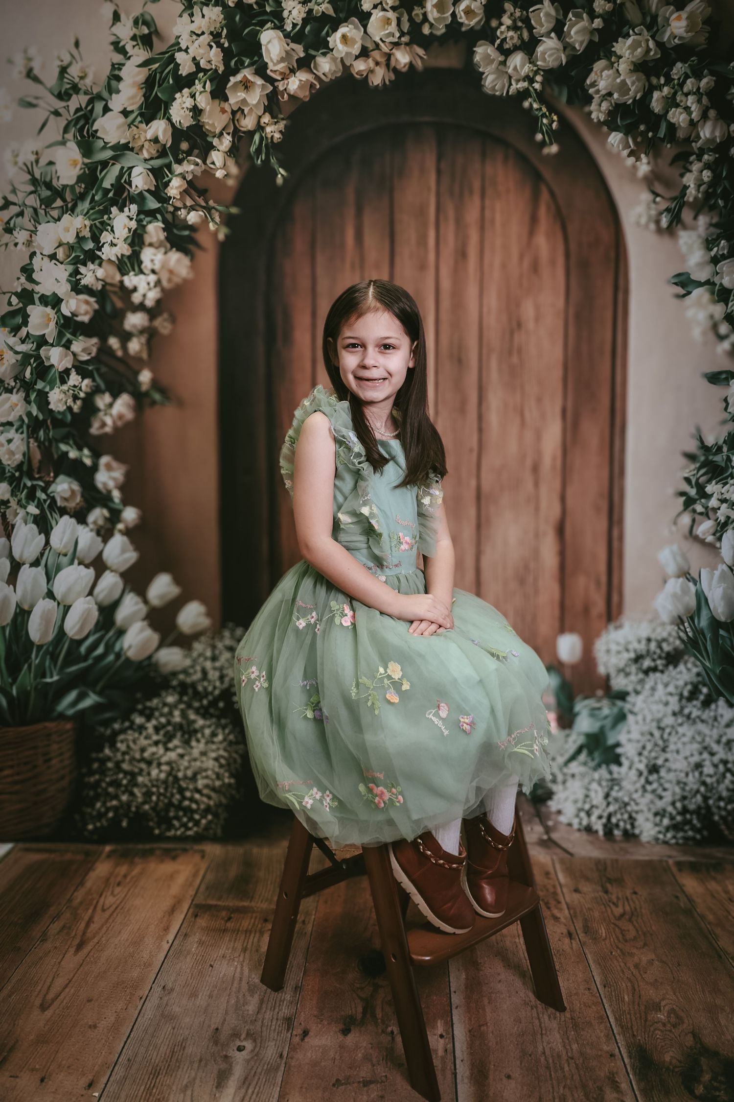 A smiling girl in a green floral tulle dress sits on a stool before a wooden door adorned with white flowers.