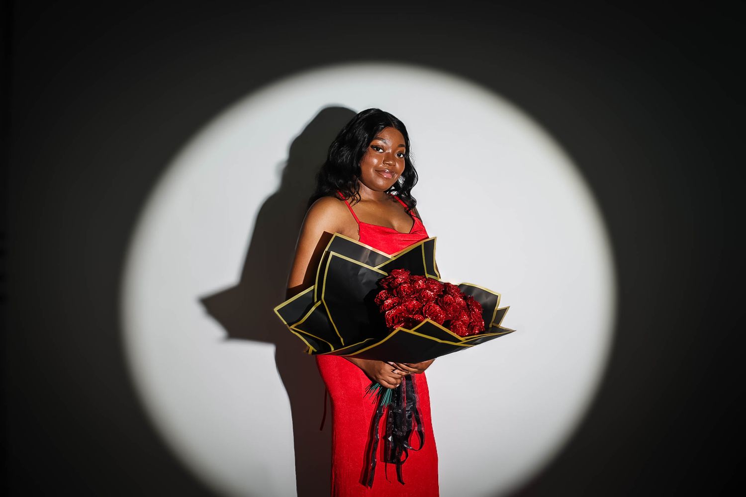 Woman in red dress holding bouquet of red roses under spotlight against white wall.