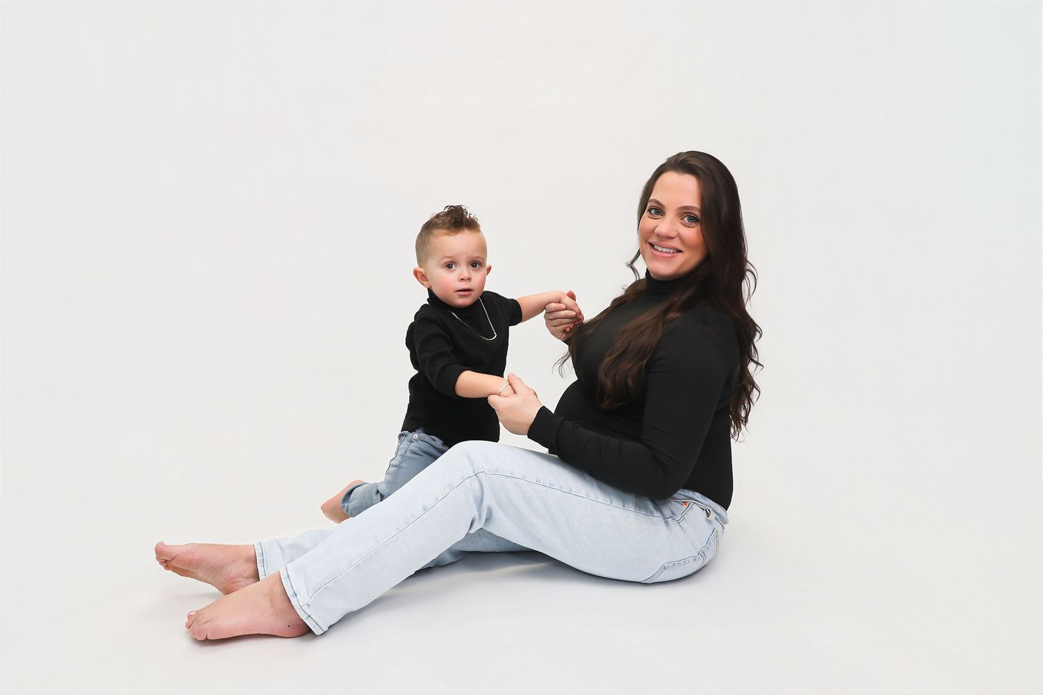 A smiling woman in black top and jeans sits on white background holding a toddler boy in matching black outfit.