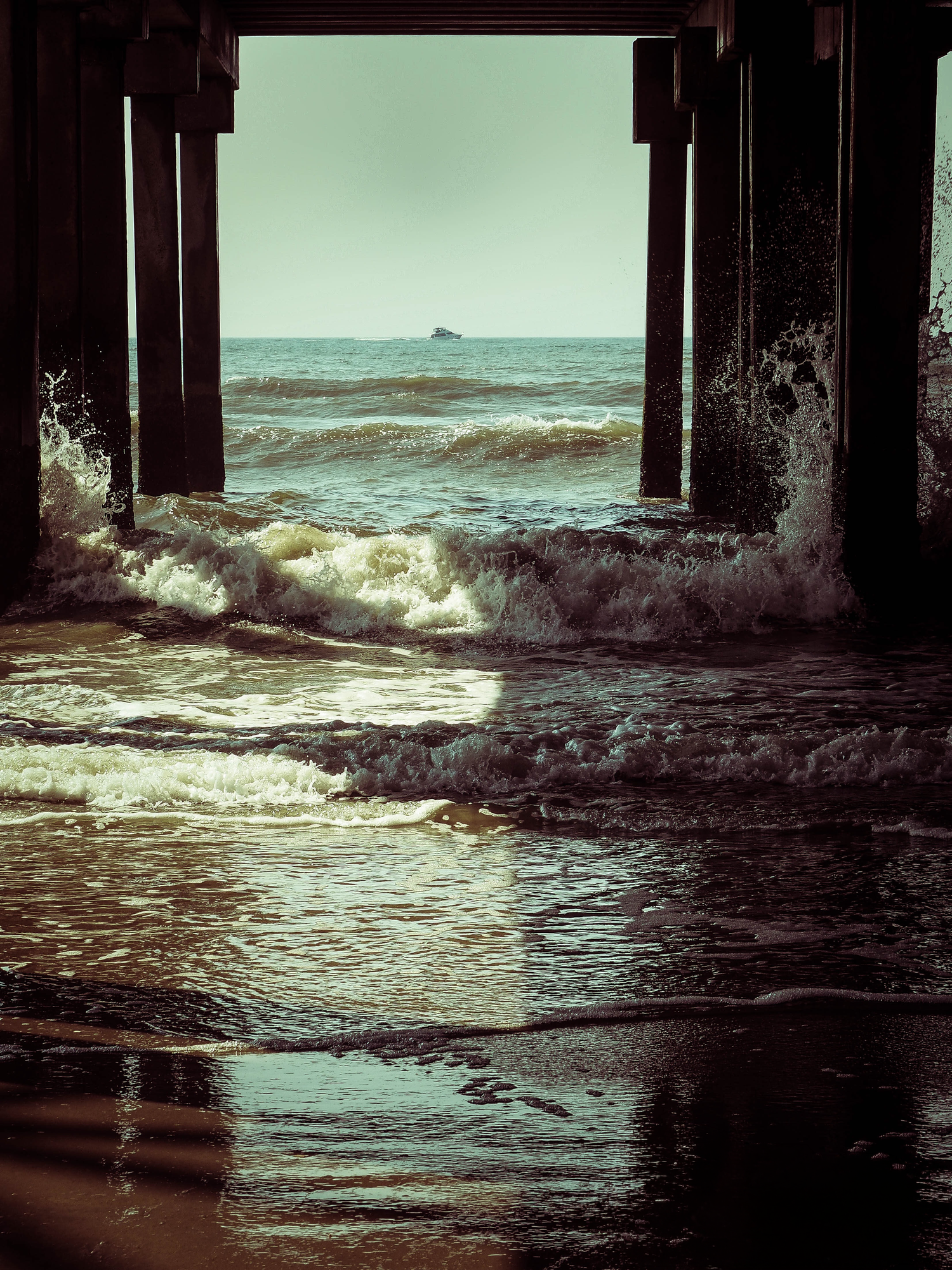 Waves crash beneath a pier with wooden pillars, reflecting golden light on wet sand, as a ship sits on the horizon.