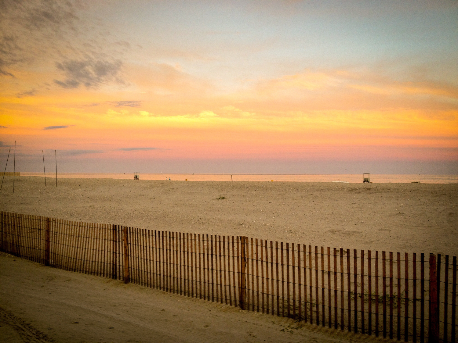 Golden sunset over a serene sandy beach with a wooden snow fence in the foreground and calm ocean horizon.