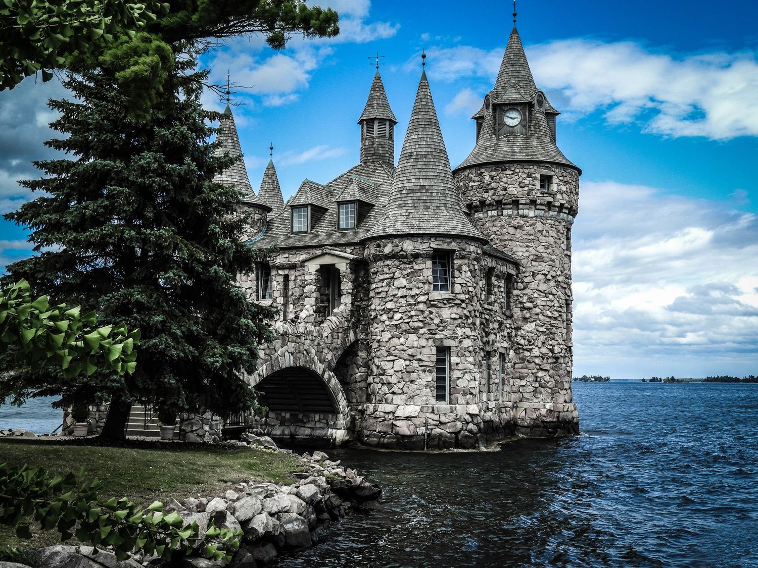 Stone castle with turrets and clock tower sits on a rocky shoreline surrounded by water under a dramatic cloudy sky.