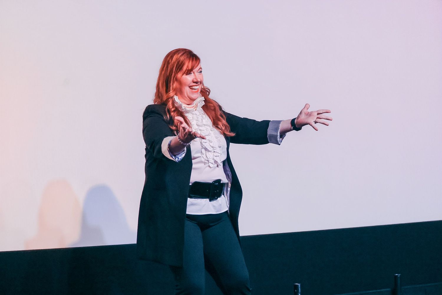 Woman with red hair gesturing expressively on stage during a presentation, wearing a dark blazer and white blouse.