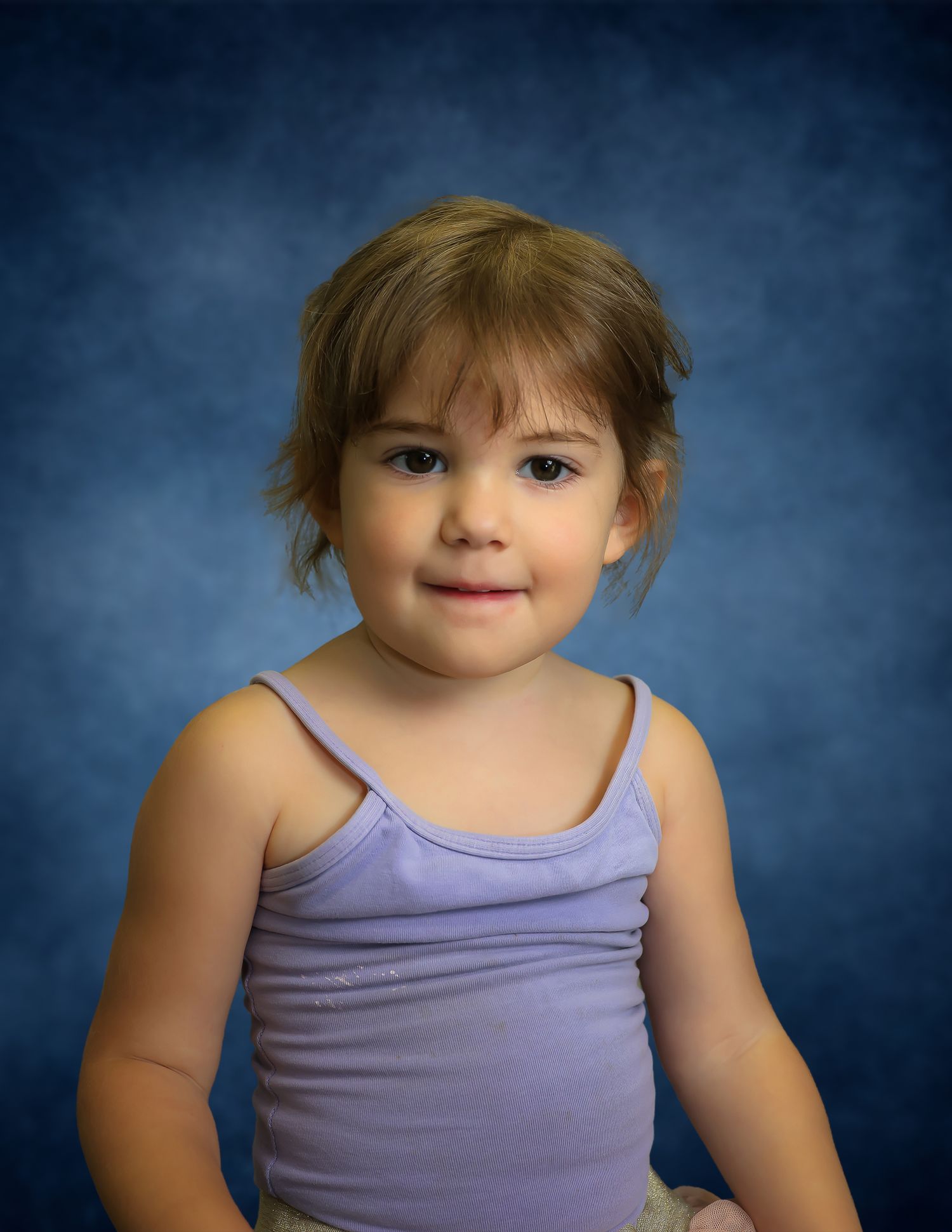 Young toddler girl with brown hair smiling in a purple tank top against a blue studio background.