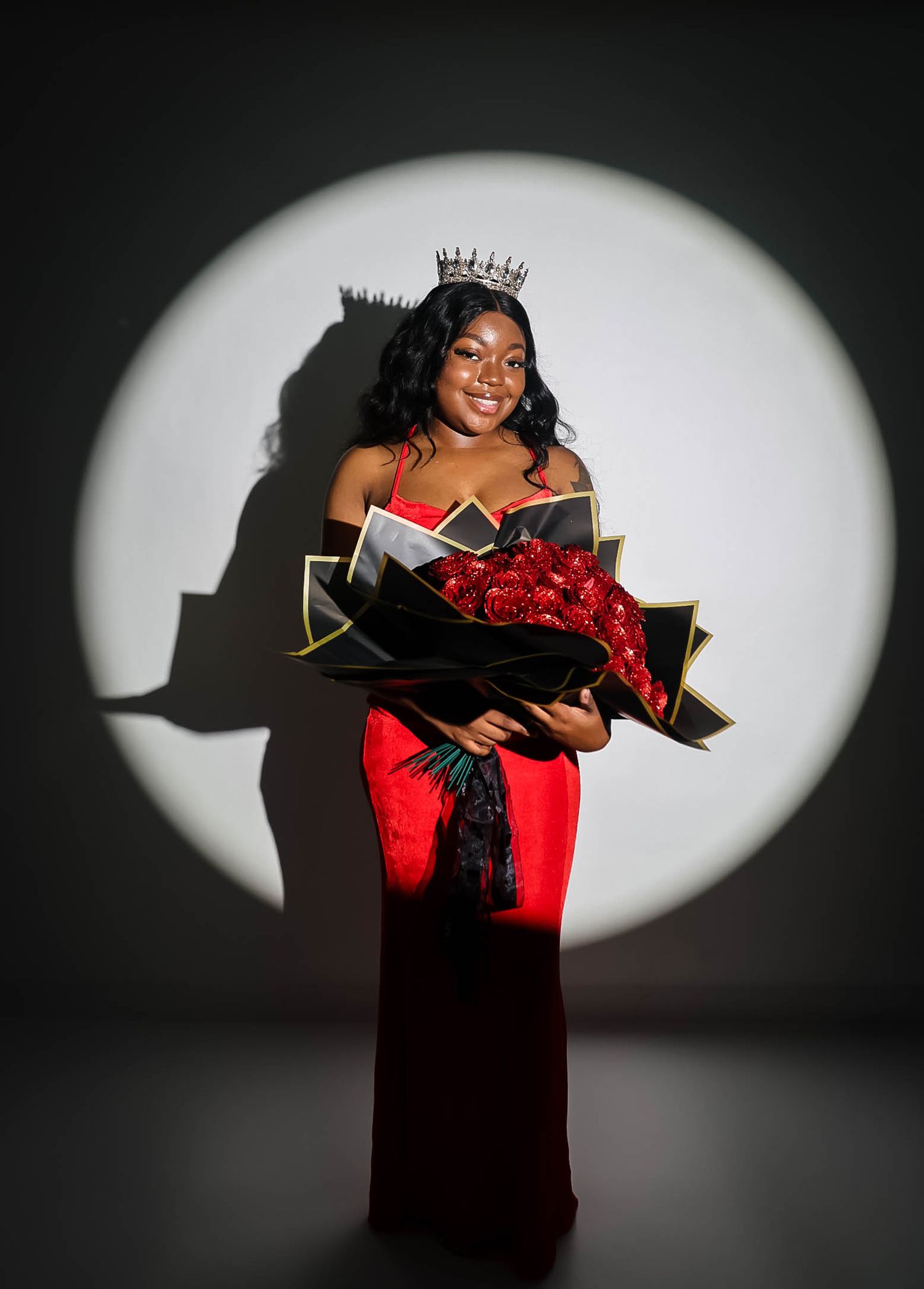 A young woman in a red gown and crown holds red roses bouquet, posed in a spotlight, beauty queen pageant portrait.