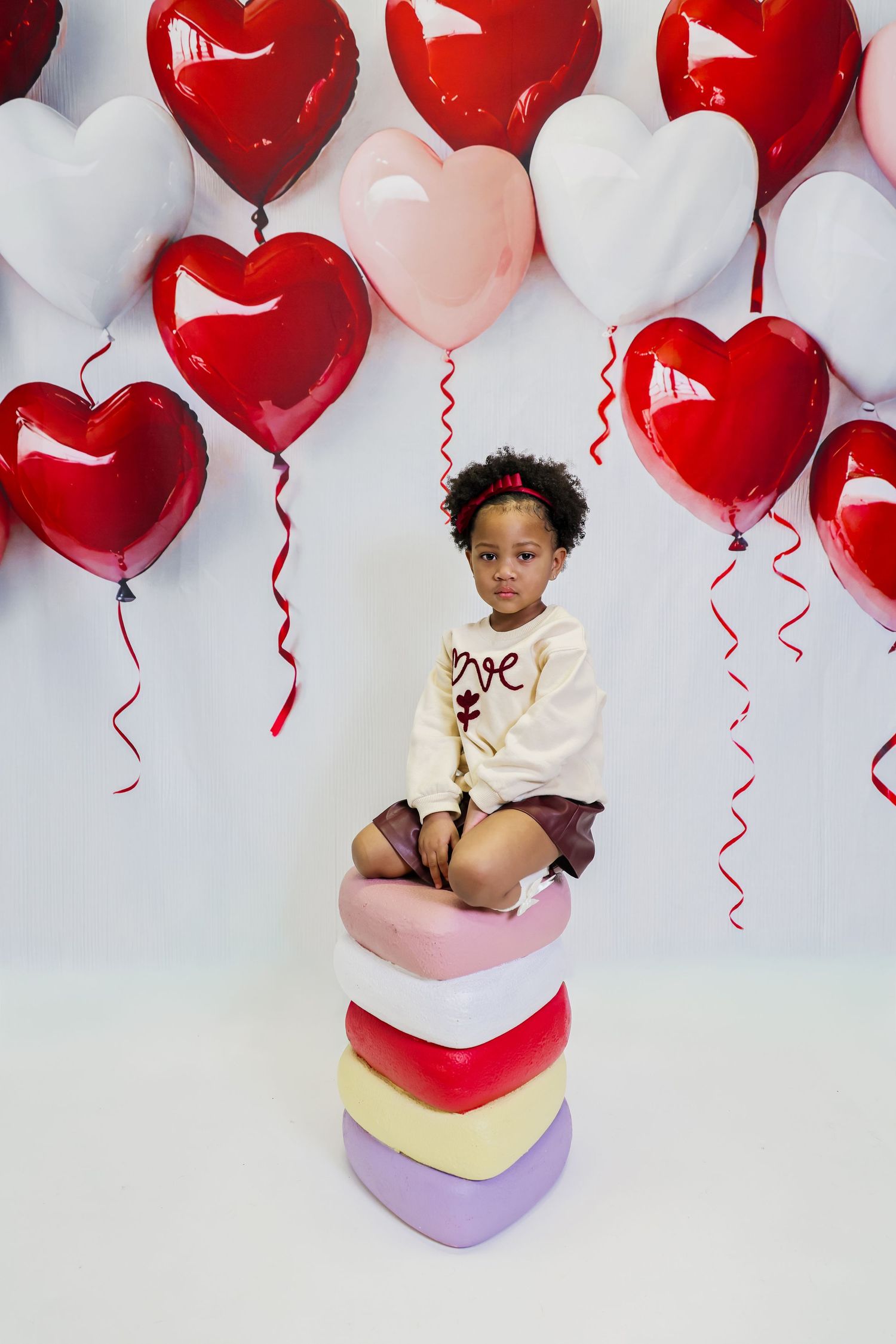 Young girl sitting on stacked heart boxes surrounded by red, pink, and white heart-shaped balloons for Valentine's Day.