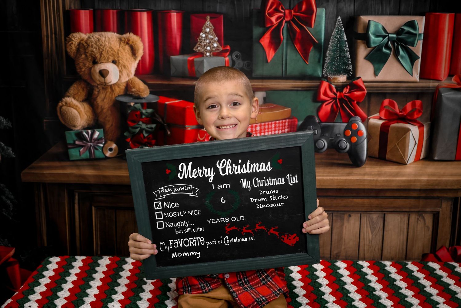 Young boy smiling and holding a Christmas chalkboard sign surrounded by festive gifts and holiday decorations.
