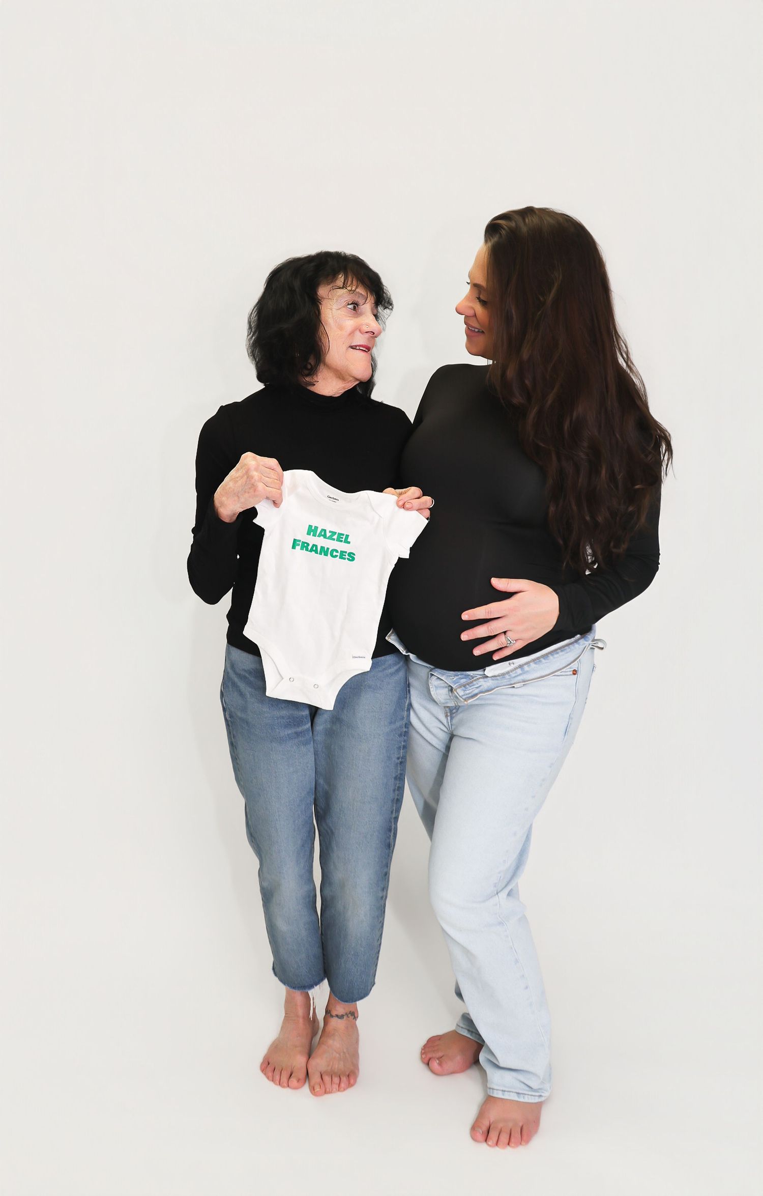 Two women holding a baby onesie reading Hazel Frances, one visibly pregnant, sharing a joyful moment together.