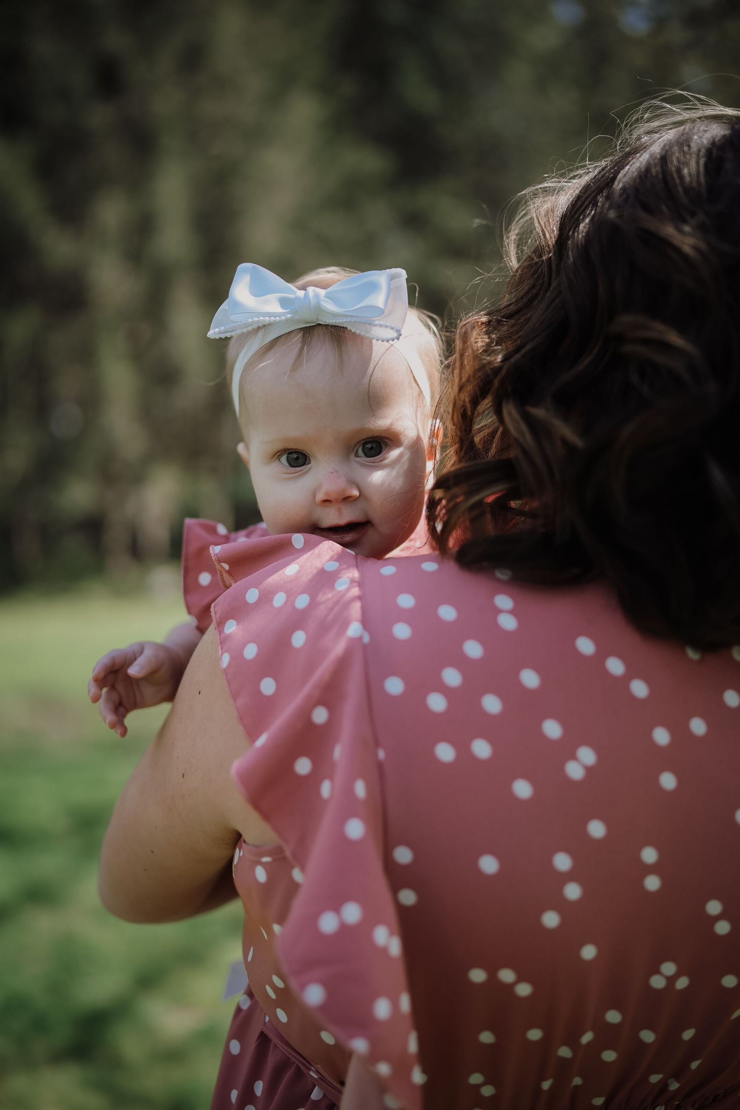 A baby girl wearing a pink polka dot dress and white bow headband being held by a curly-haired woman outdoors.
