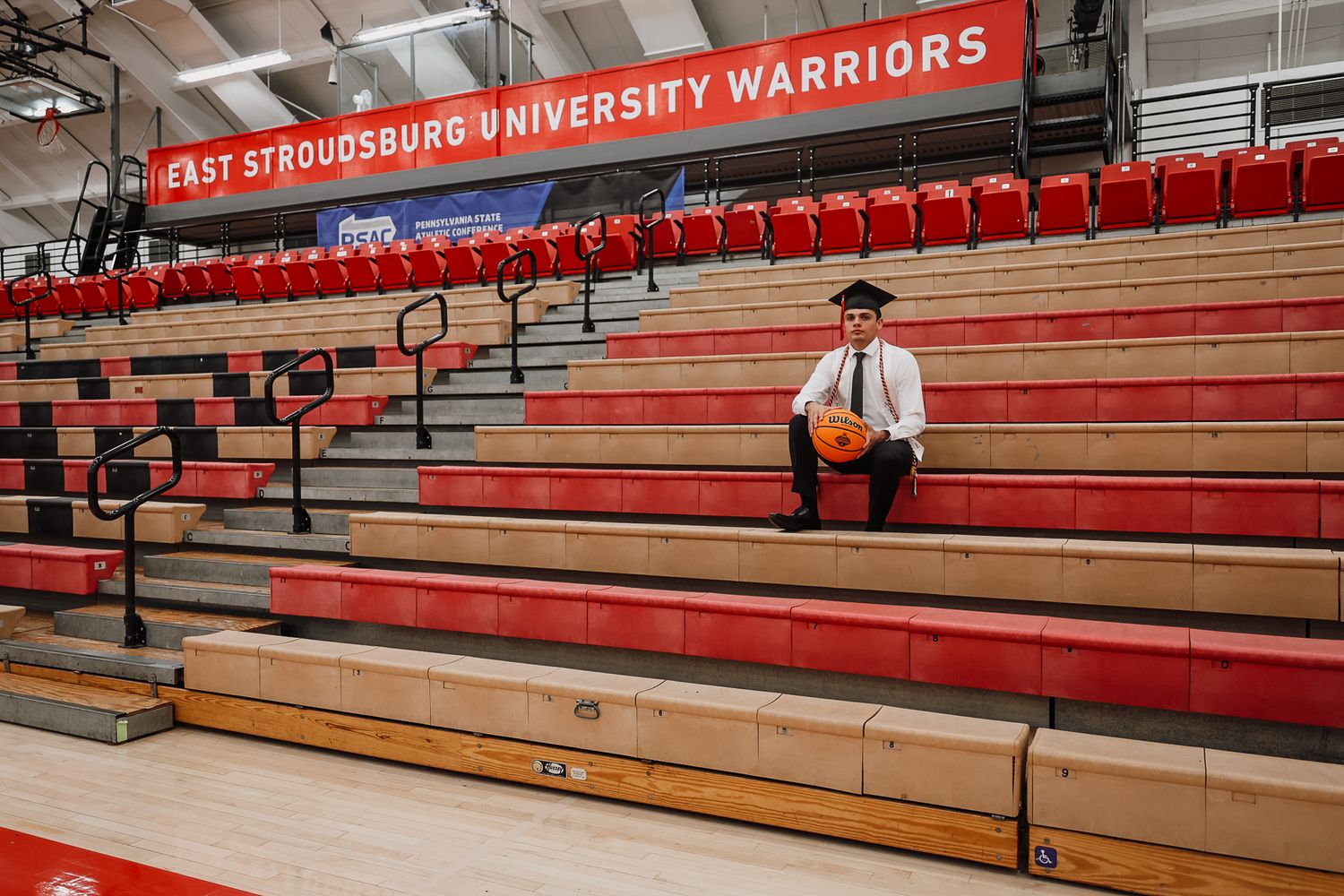 Graduate in cap and gown sits alone in East Stroudsburg University Warriors gymnasium bleachers holding a basketball.