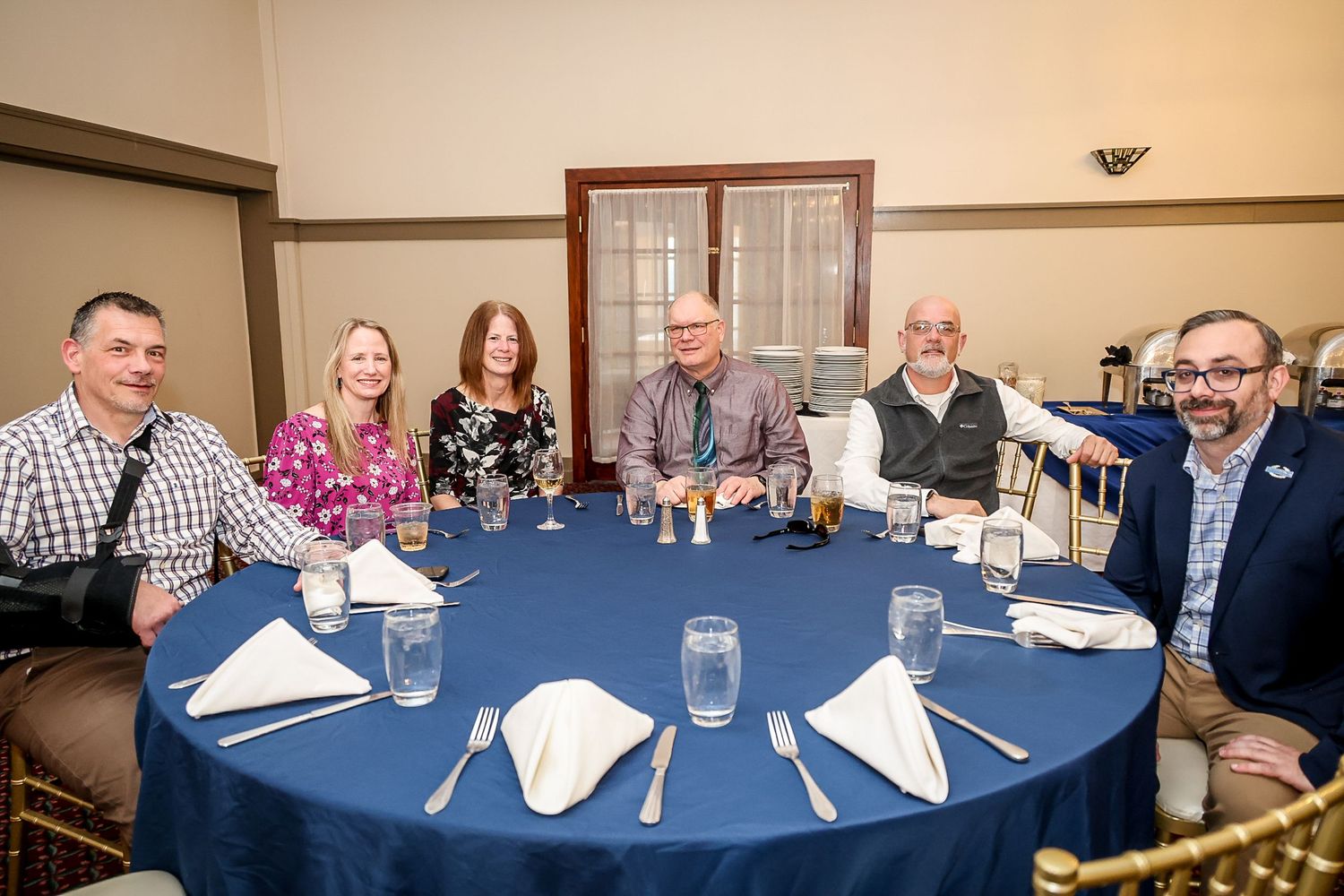 Six people smile at a round banquet table with blue tablecloth, set for a formal dinner event.