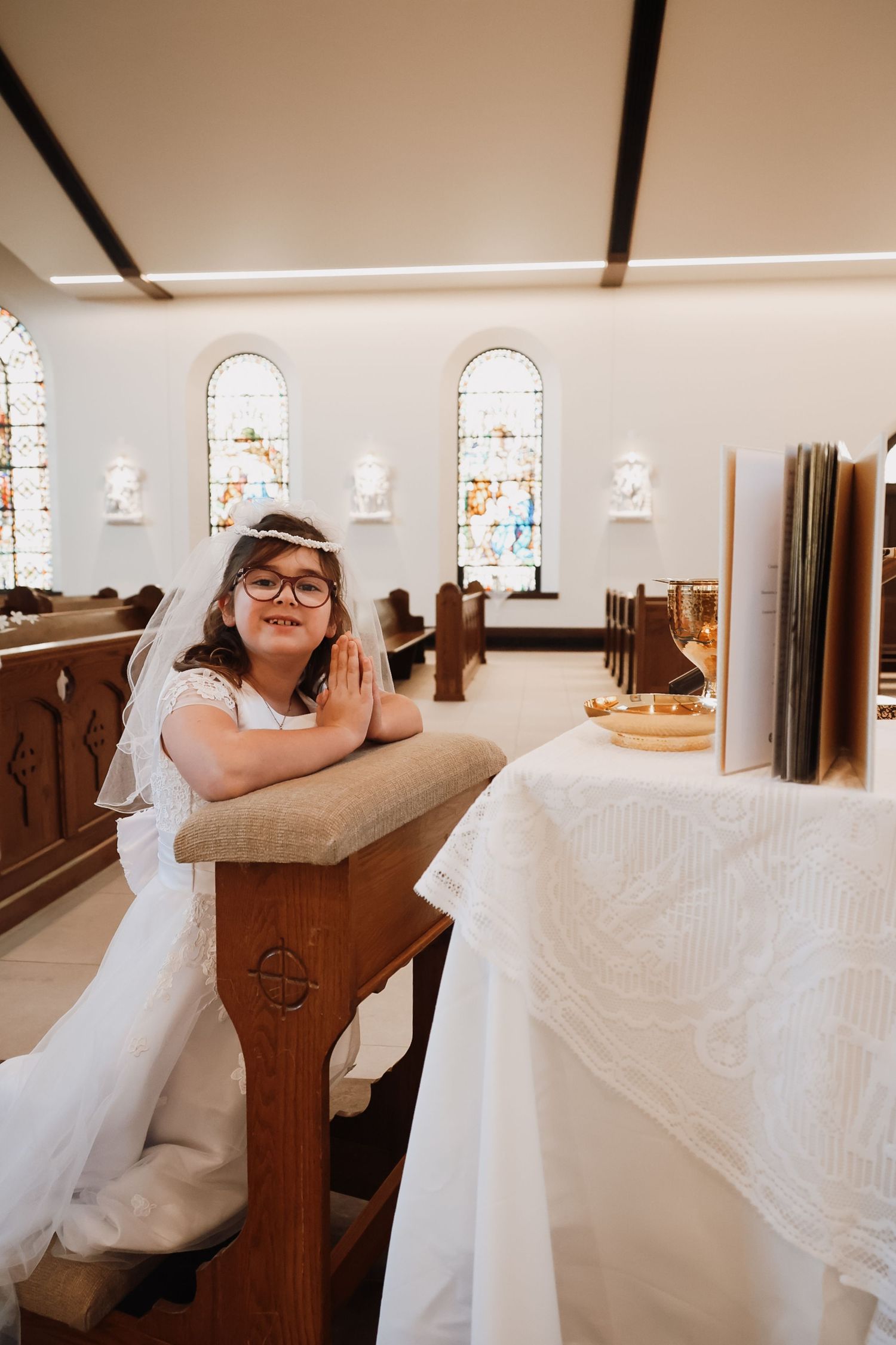 Young girl in white dress and veil praying at church pew during First Communion ceremony.