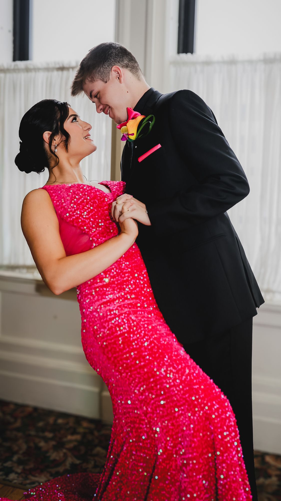 A couple in formal attire, woman in red sequin gown and man in black tuxedo, dancing together at prom.