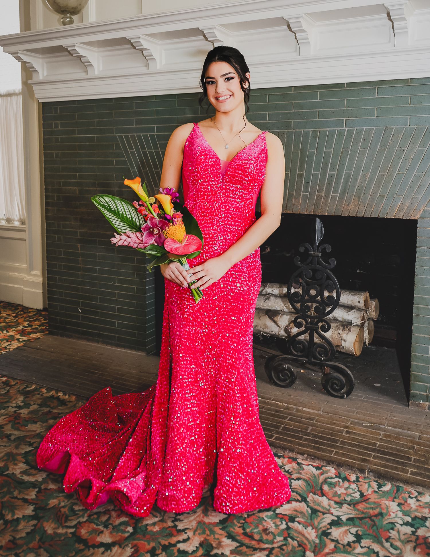 Woman in a red sequin mermaid gown holding a tropical bouquet, posing in front of a fireplace.