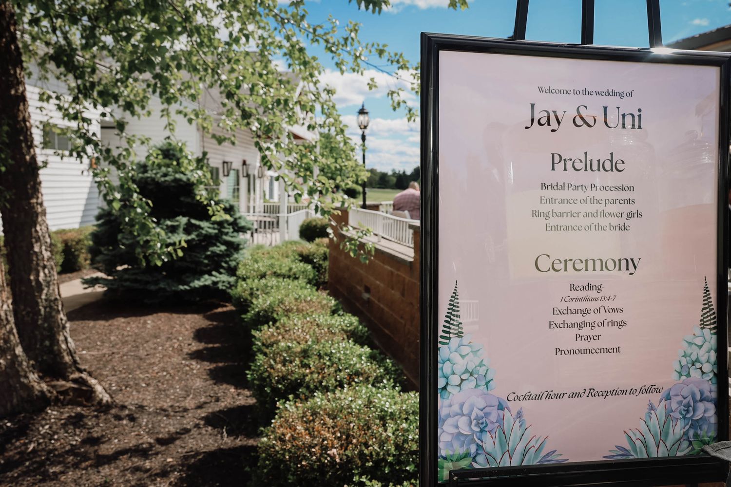 Elegant outdoor wedding sign for Jay and Uni displaying ceremony order including prelude and vows amid lush garden backdrop.