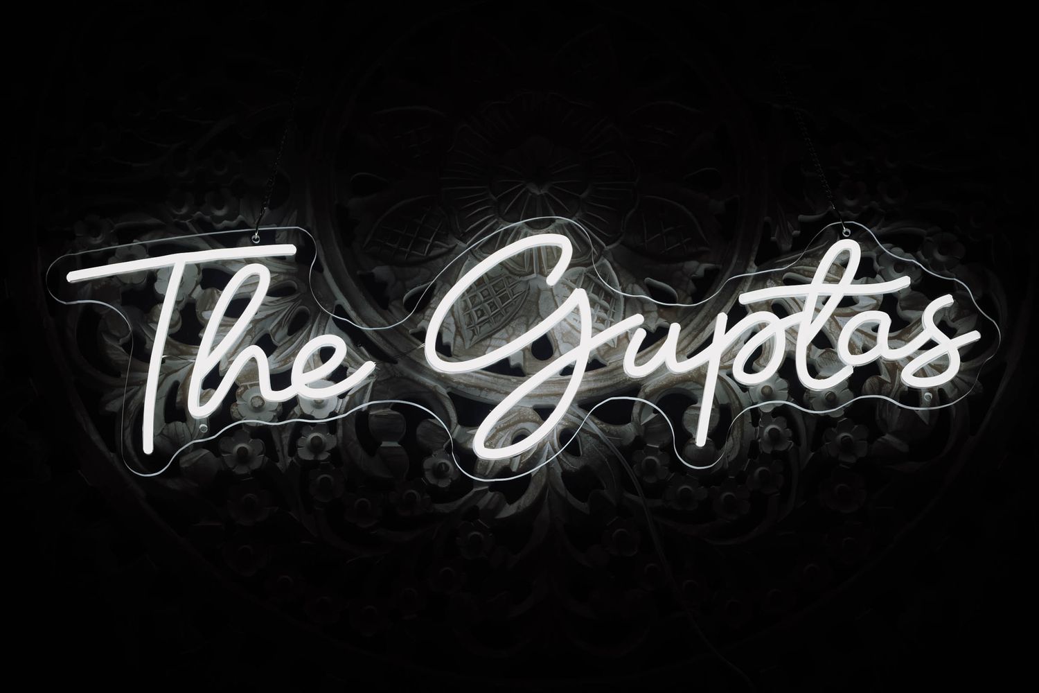 White neon sign reading 'The Guptos' in cursive script against a dark black background.