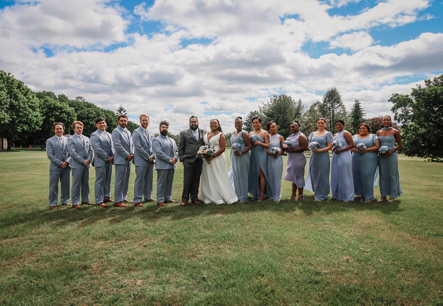 Wedding party poses outdoors on green lawn, groomsmen in gray suits and bridesmaids in dusty blue dresses flanking bride and groom.