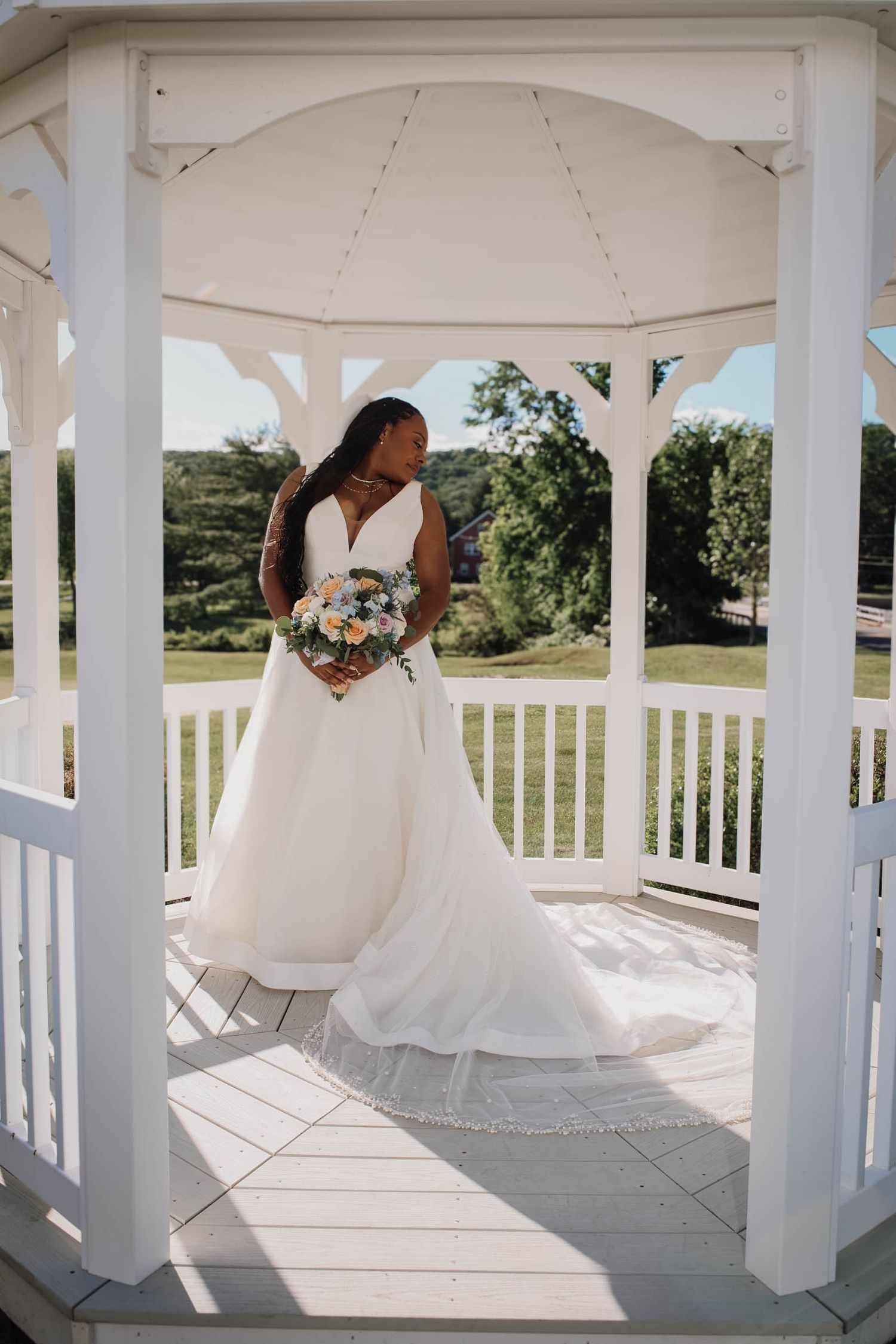 Bride in white gown holding colorful bouquet stands in white gazebo surrounded by lush greenery on sunny wedding day.