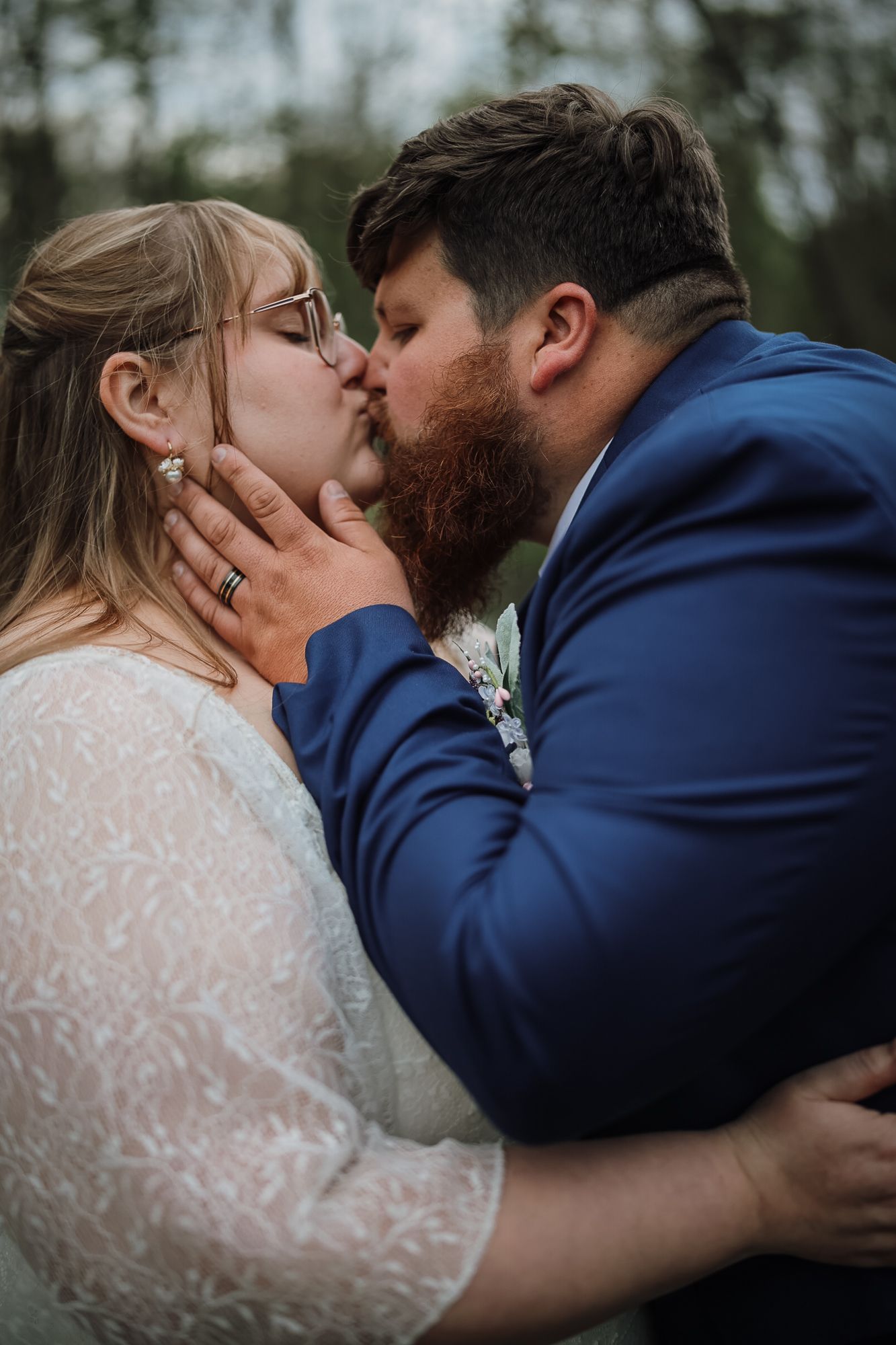 Bride and groom sharing a romantic first kiss, bride in lace gown with glasses, groom in navy blue suit with red beard.