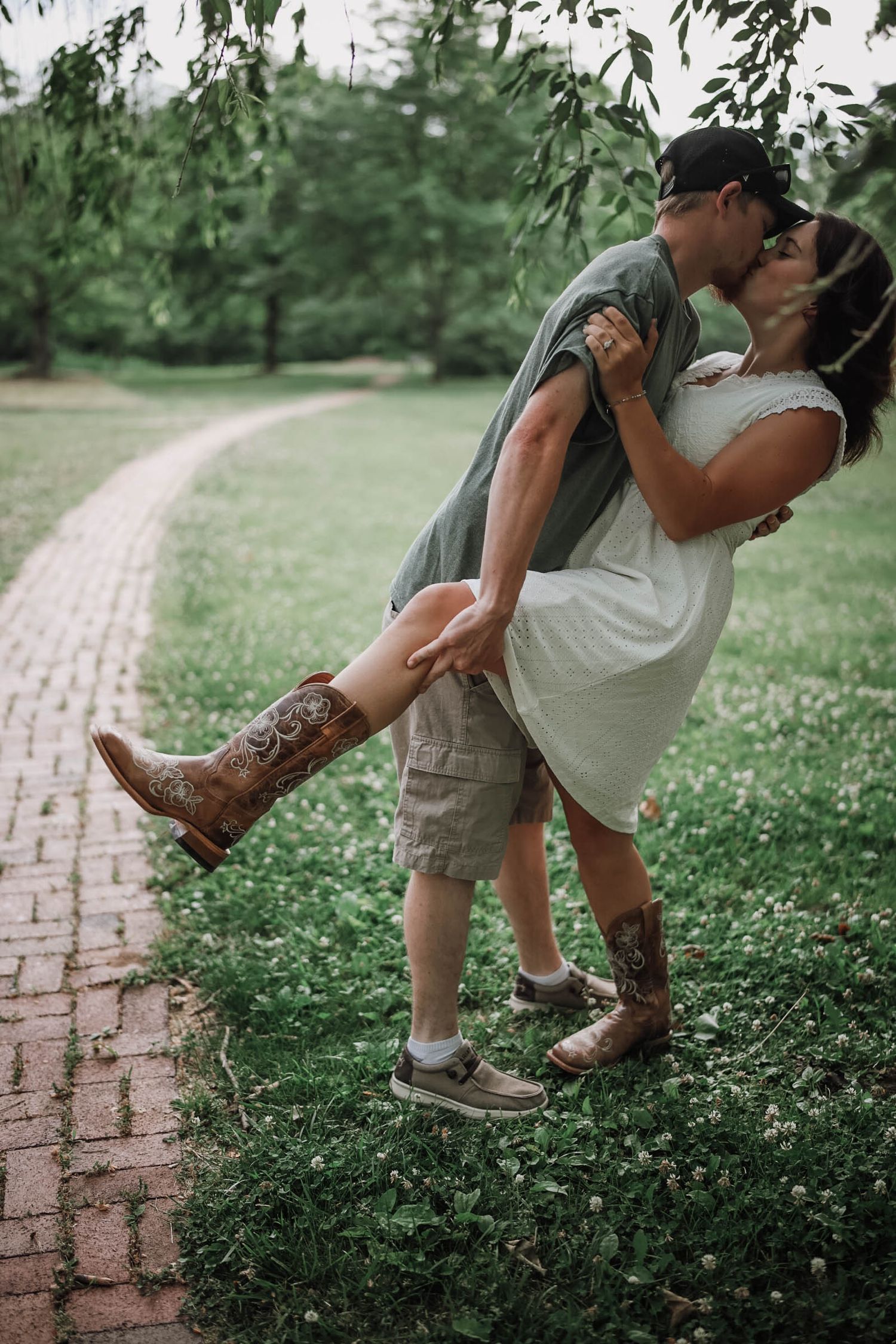 Man lifting woman for a romantic kiss on a brick pathway surrounded by green grass and weeping willow trees.