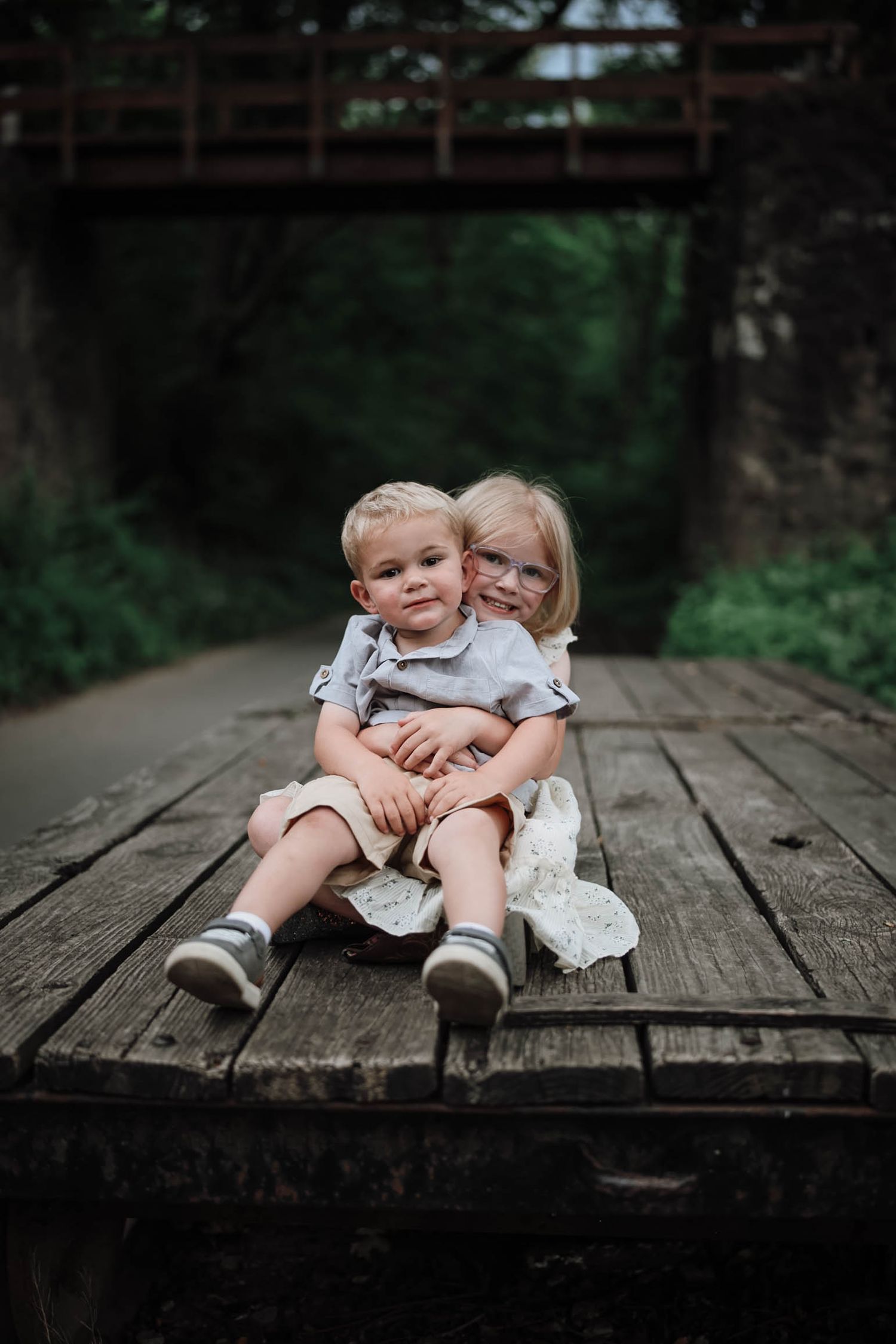 Two young siblings sitting on a wooden bridge, older girl hugging younger boy, surrounded by lush greenery.