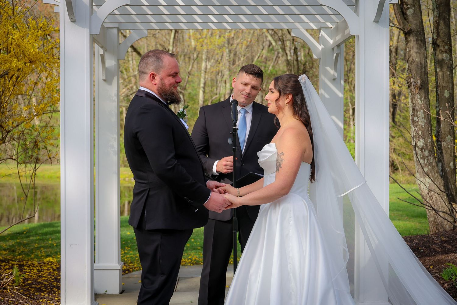 Couple exchanges vows under white garden arbor with officiant, bride in white gown and veil, groom in black tuxedo.