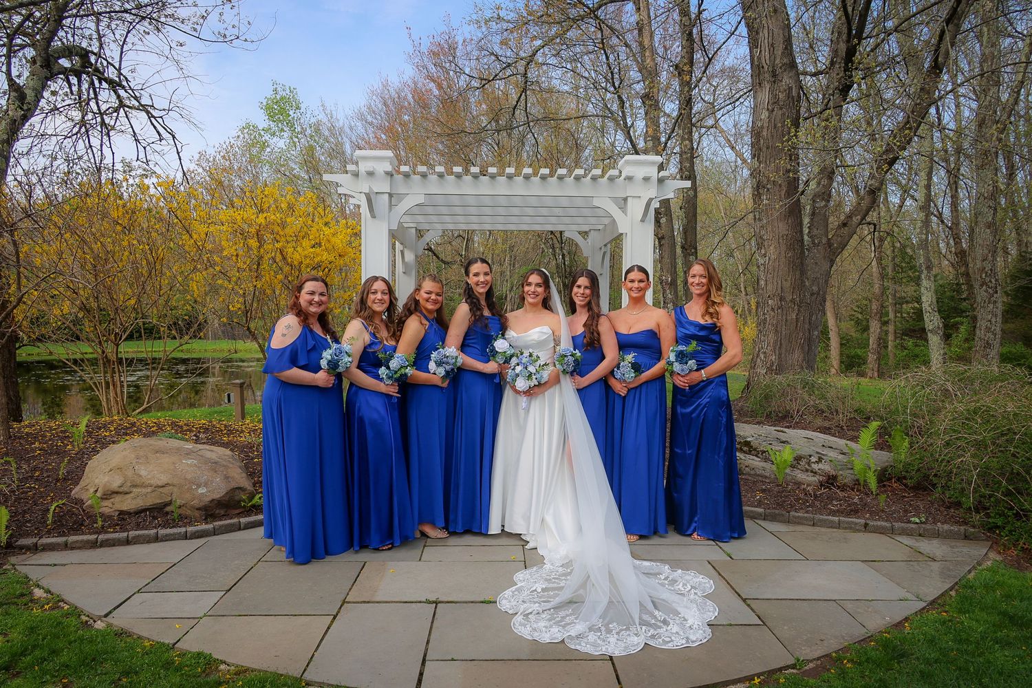 Bride in white gown poses with six bridesmaids in royal blue dresses holding bouquets before a white pergola outdoors.
