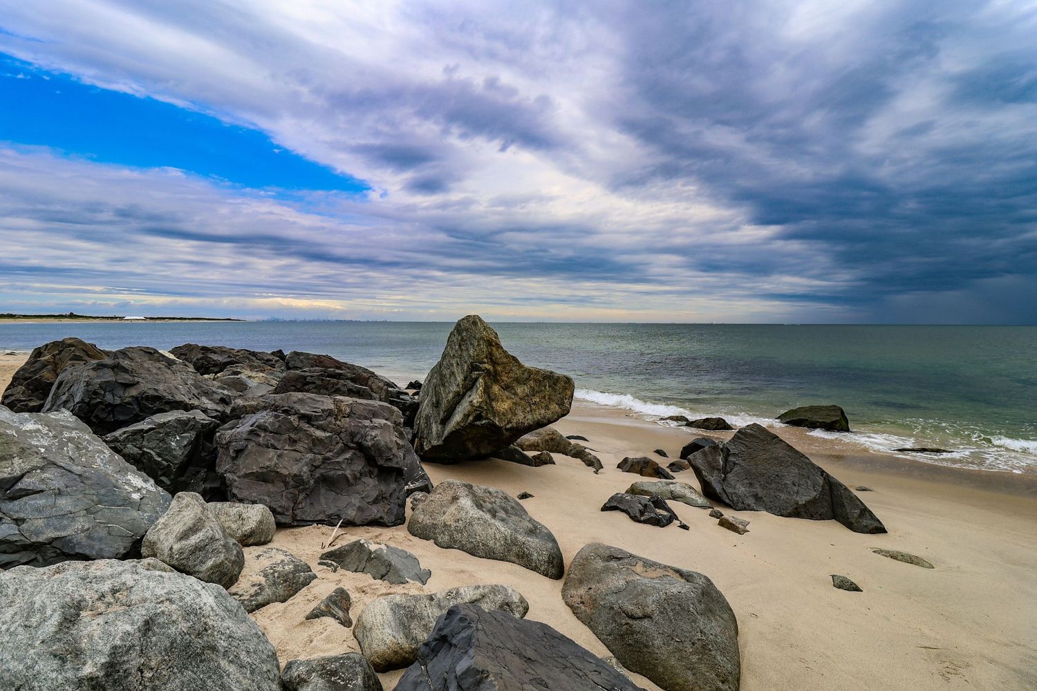 Rocky shoreline with large boulders on sandy beach under dramatic cloudy sky with calm ocean waves in background.