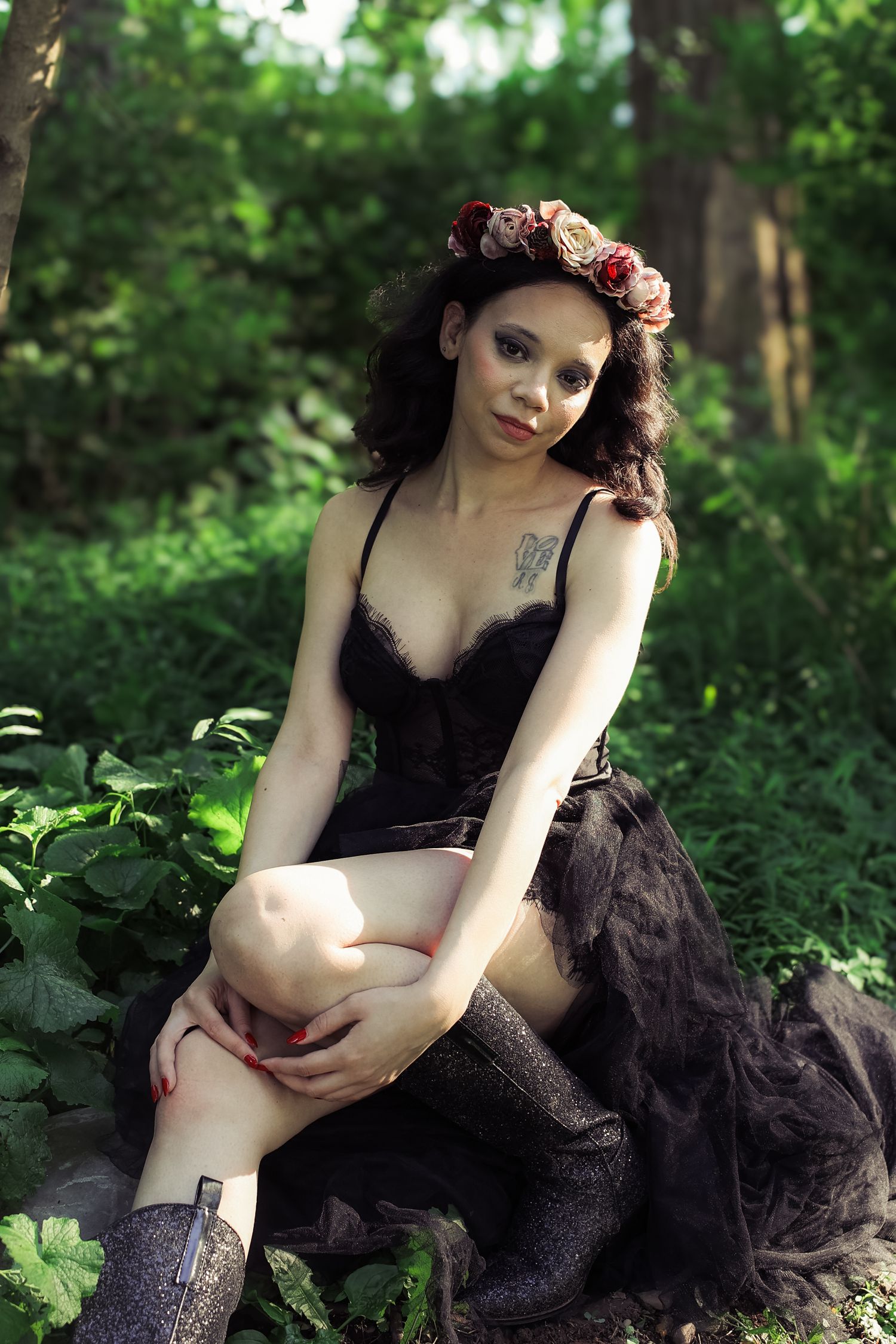 A figure in a black dress poses gracefully in a lush forest setting with flowers in their hair.