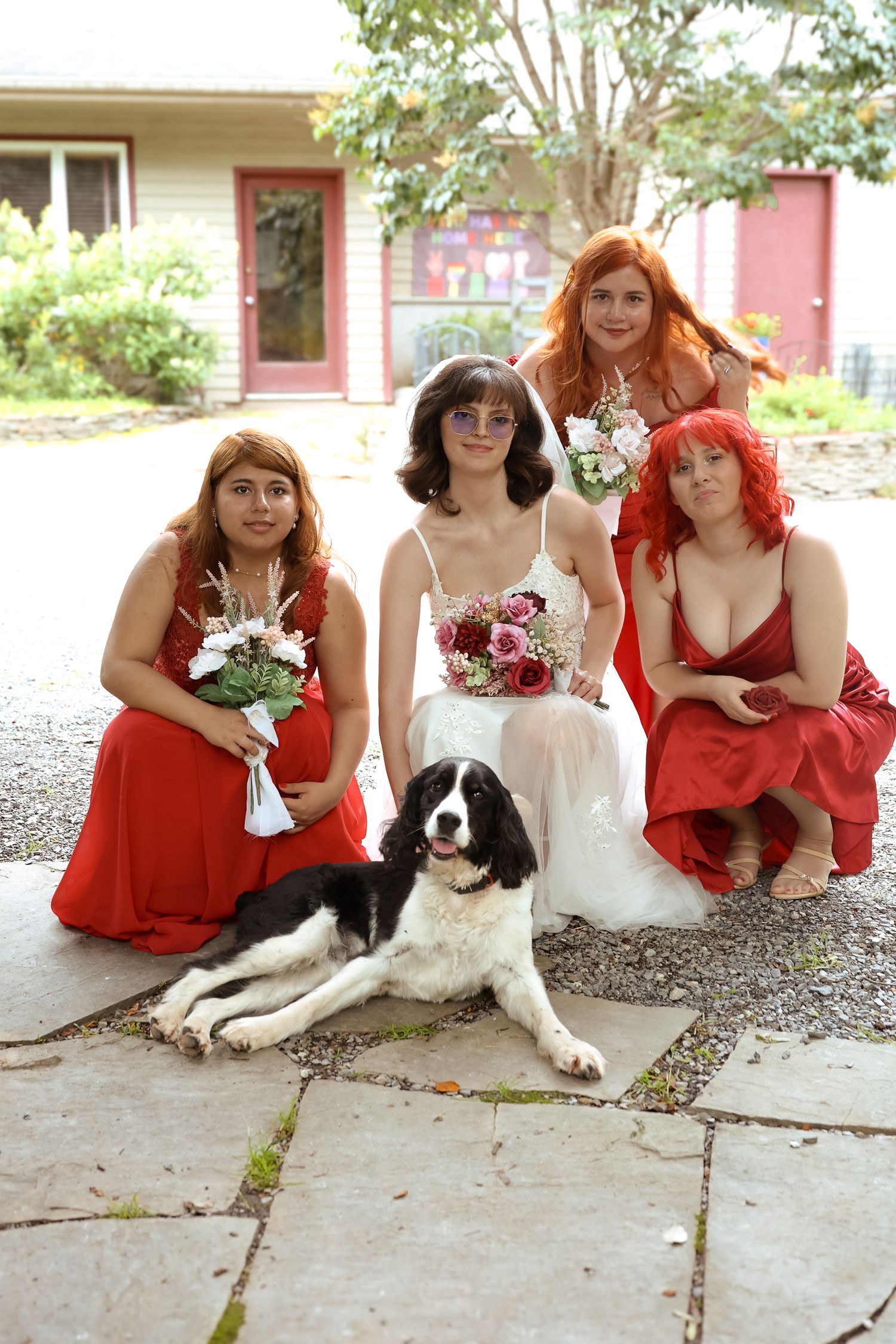 A wedding party in red dresses poses with a black and white dog on a porch.