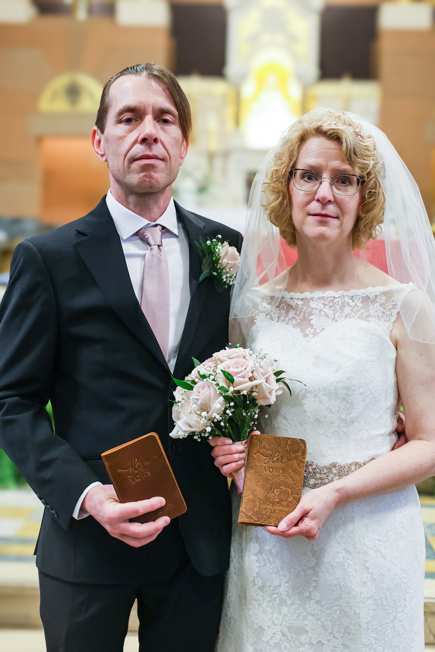 A couple stands together at their wedding ceremony holding brown leather-bound books while wearing formal attire.