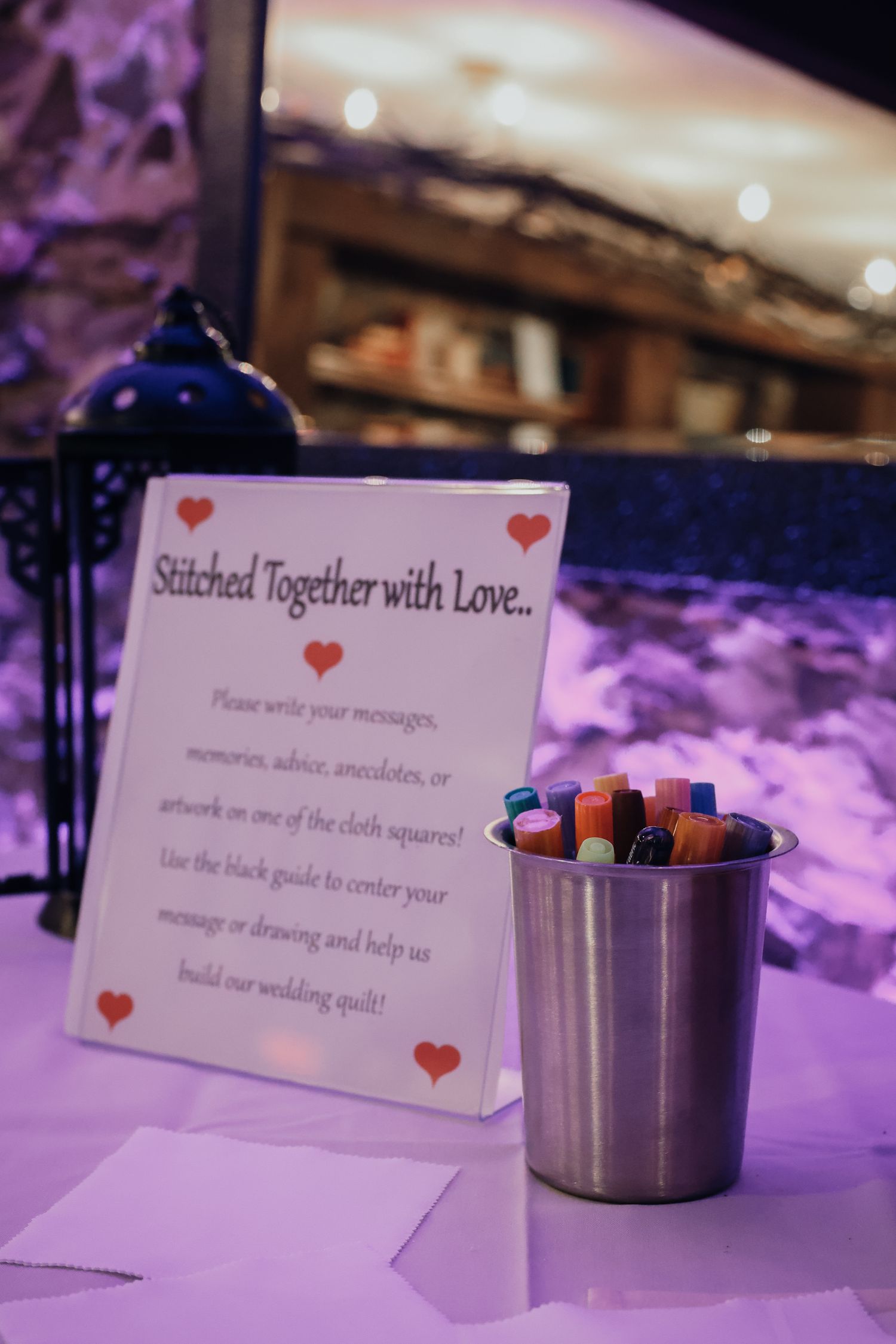 A wedding guest book sign reading 'Stitched Together with Love' with colored markers in a metal cup on a purple-lit table.