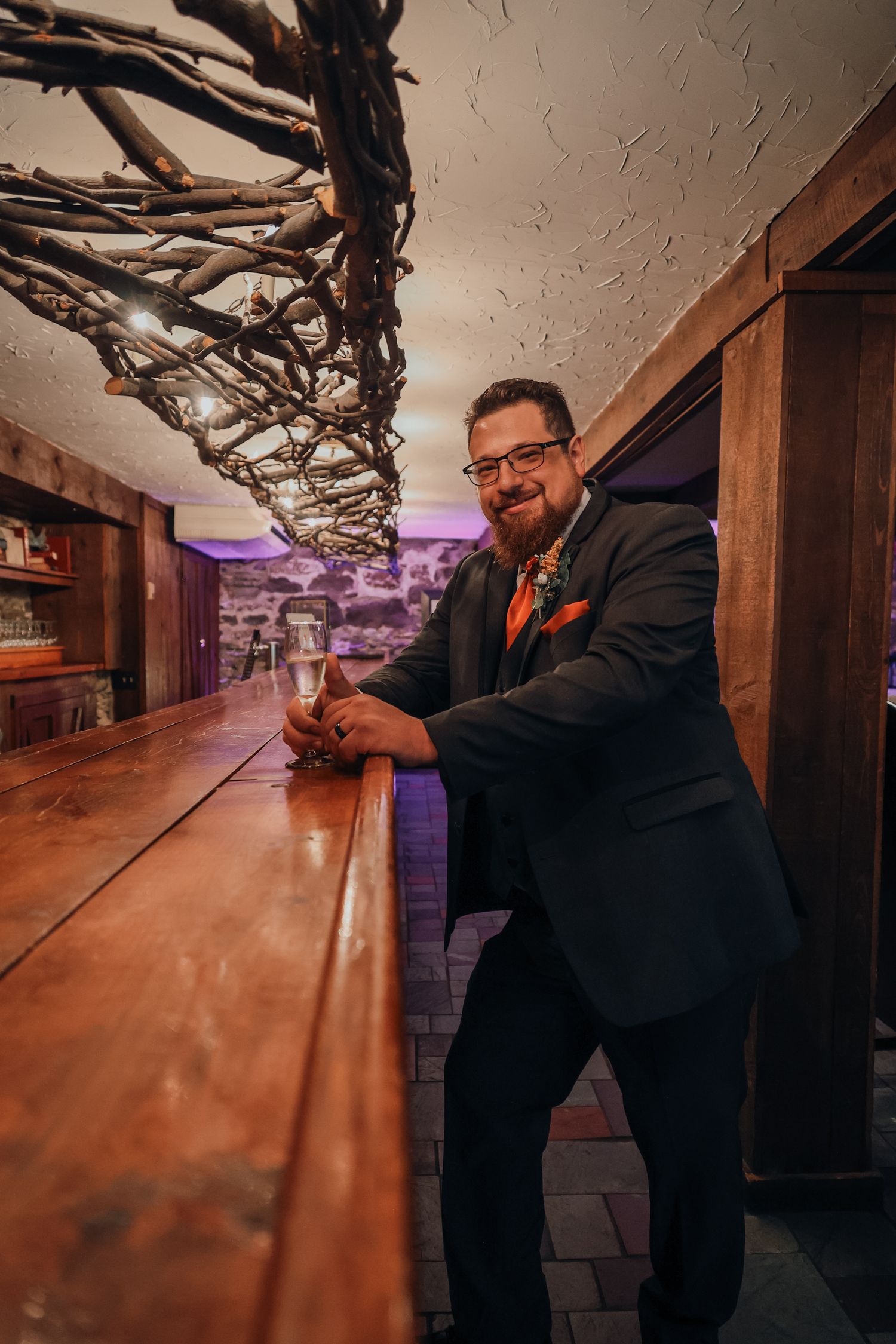 A person in a black suit and orange tie stands at a rustic wooden bar with decorative branches overhead and purple lighting.