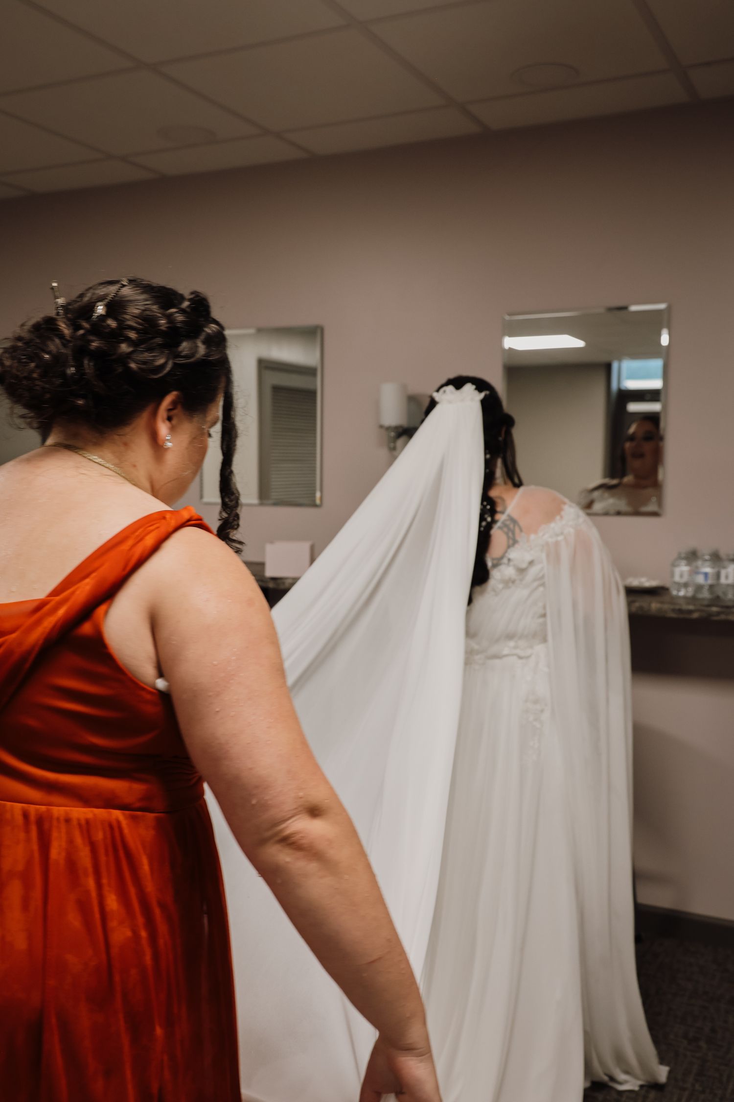 A bridesmaid in a red dress helps adjust a white wedding gown hanging on display in a dressing room.