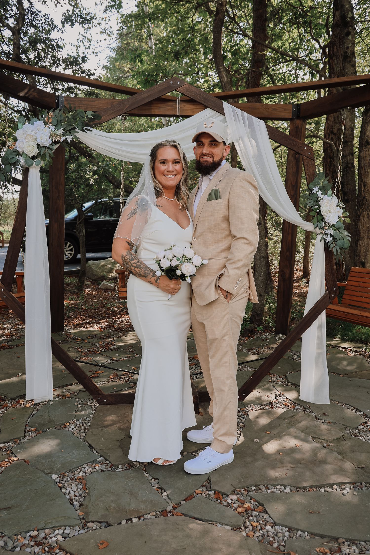A couple poses under a wooden arch decorated with white fabric and flowers at an outdoor wedding ceremony.