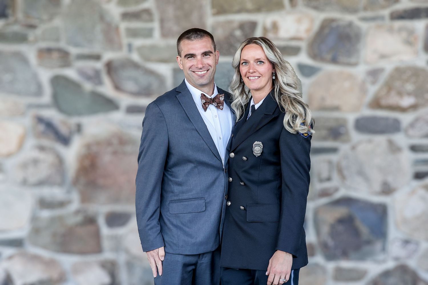 A couple in formal navy attire poses together against a rustic stone wall background.