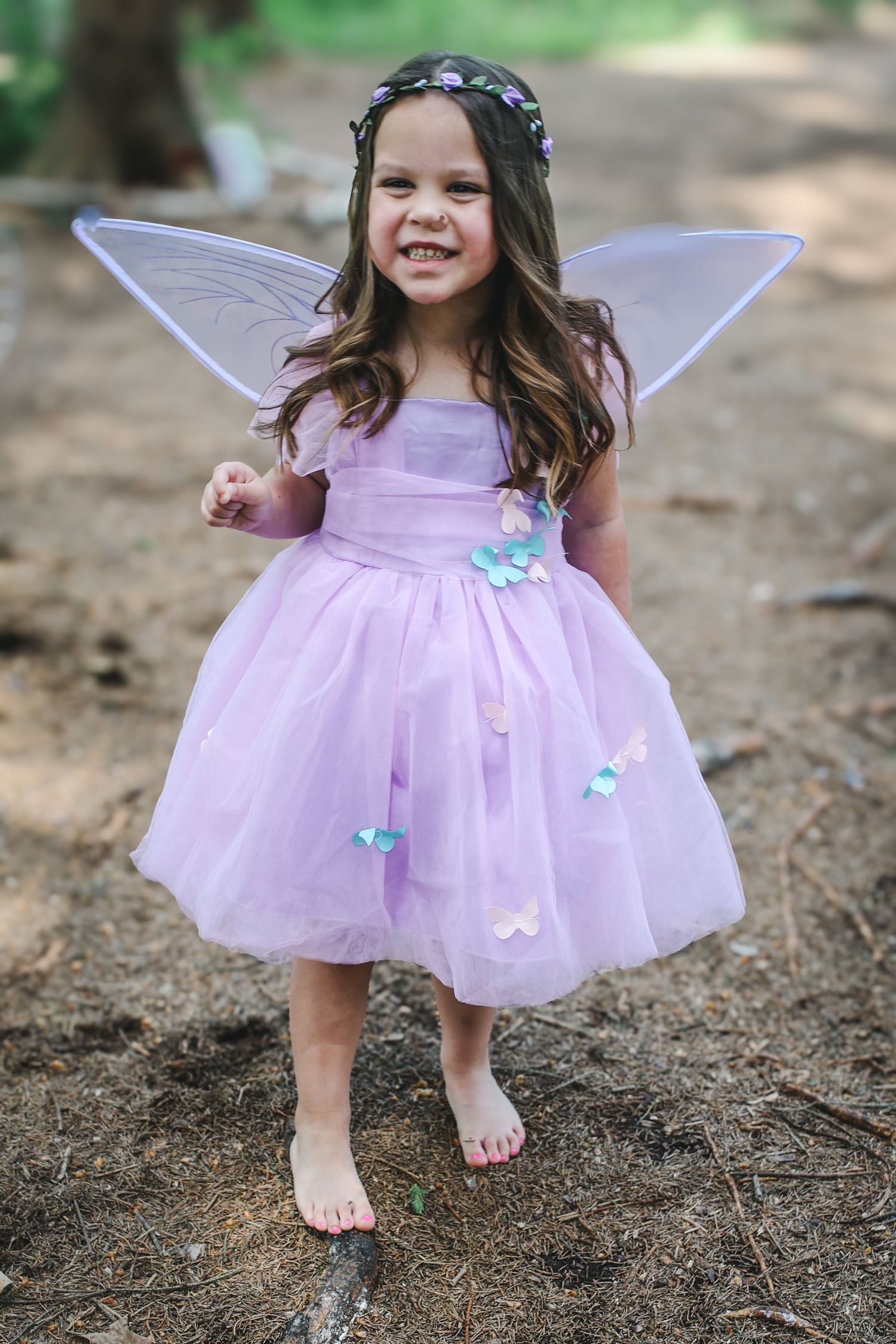 A pink fairy dress with wings and butterfly details is worn outdoors in a woodland setting.