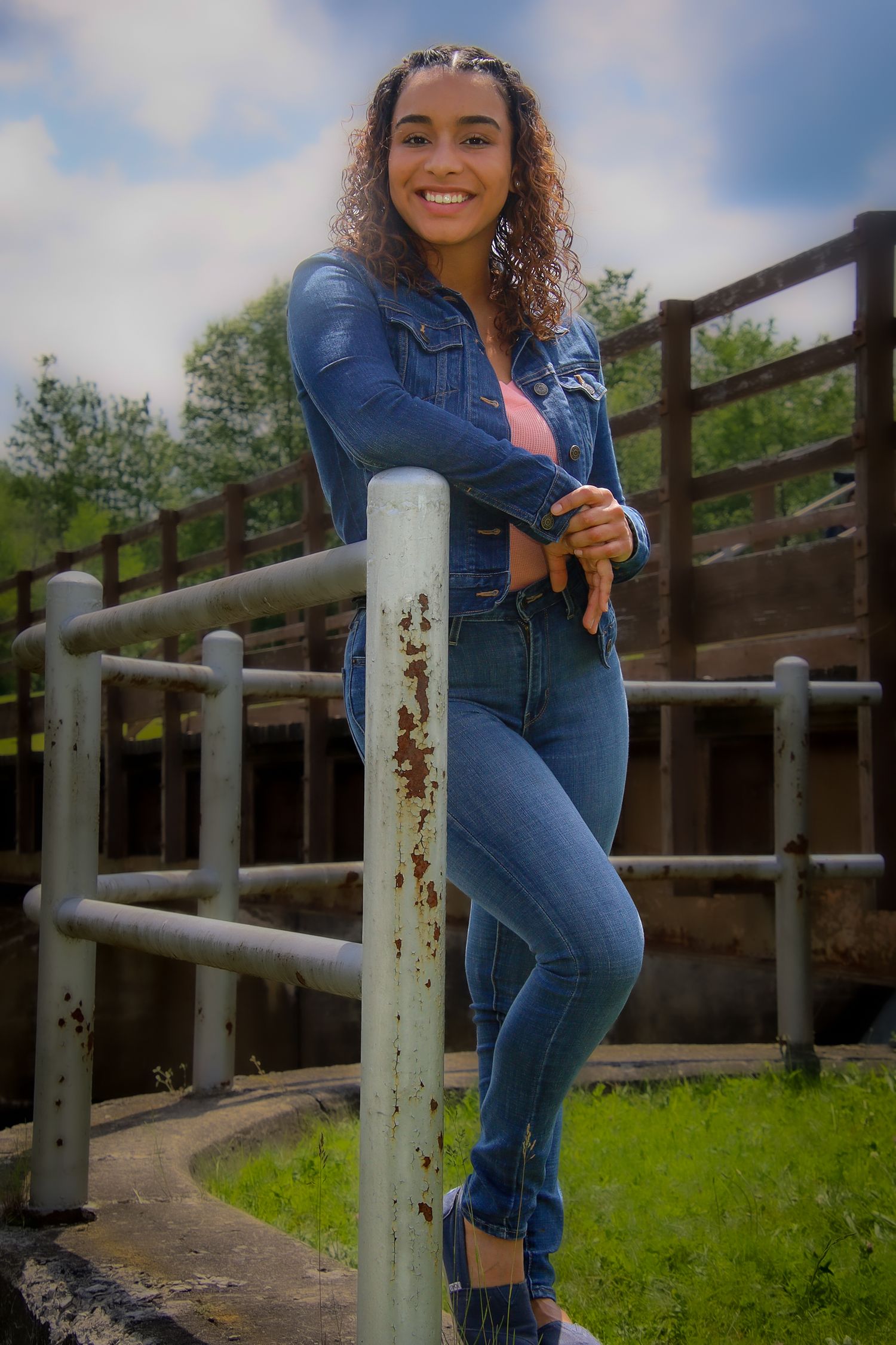 A person in denim overalls poses casually on a wooden fence railing in a rural outdoor setting.