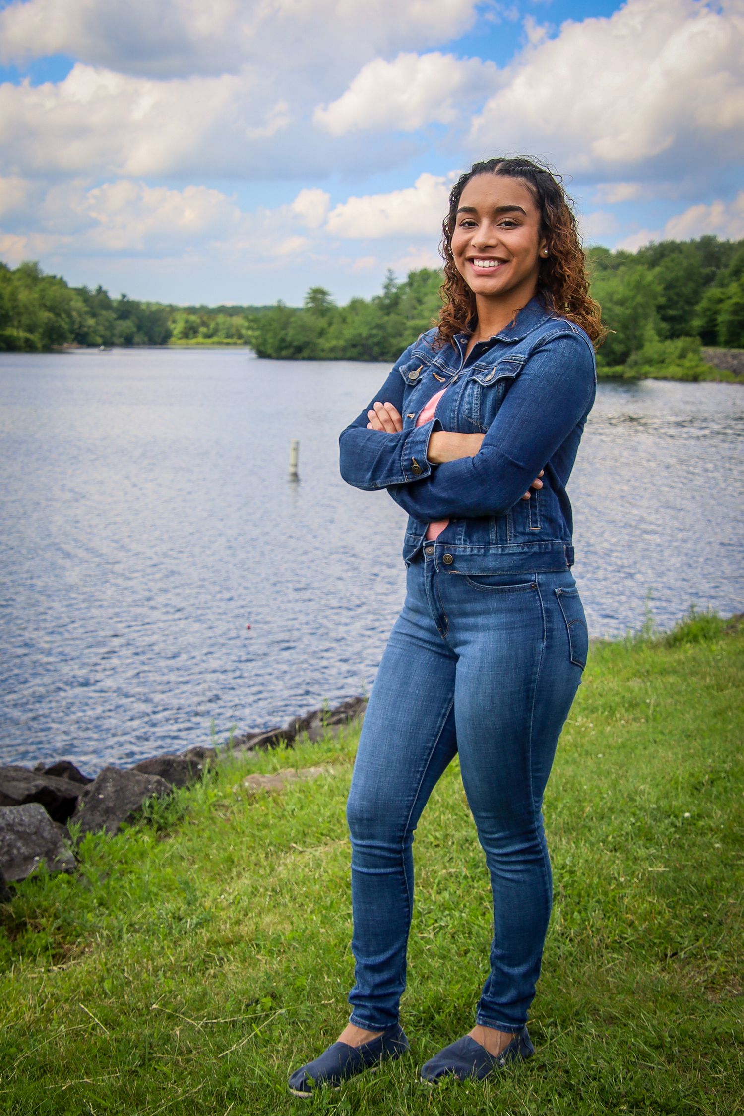 A person in denim overalls and blue shirt poses confidently by a scenic lake with green grass and cloudy skies.