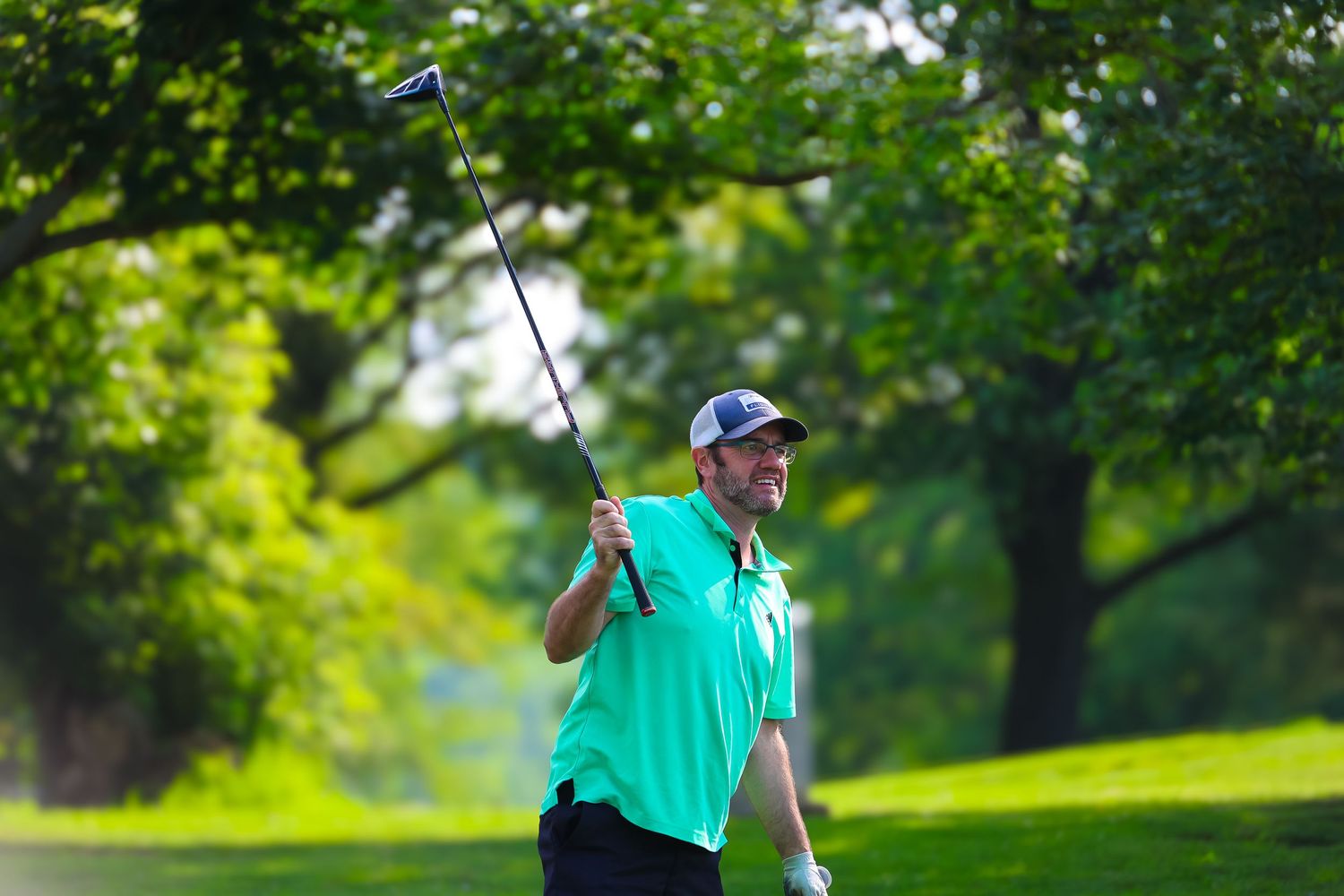 A golfer in a turquoise shirt takes a swing on a lush green golf course surrounded by trees on a sunny day.