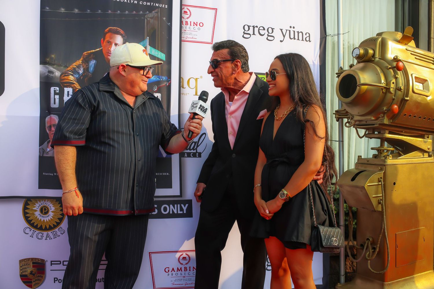 Three people interact on a red carpet event with a golden decorative prop visible in the background.