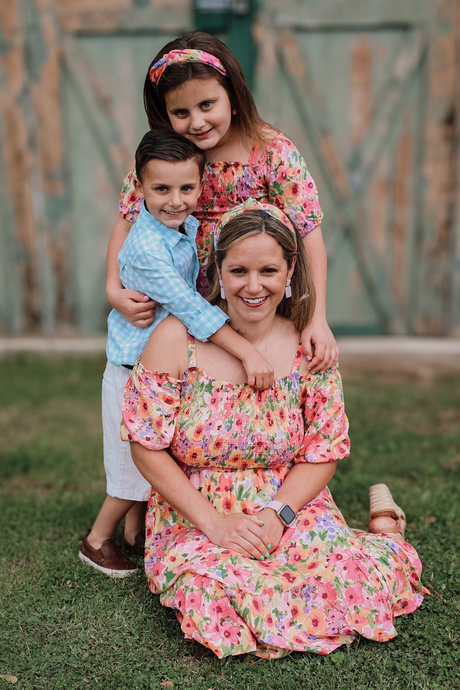 Family wearing coordinated spring outfits poses together on grass in front of rustic barn door.