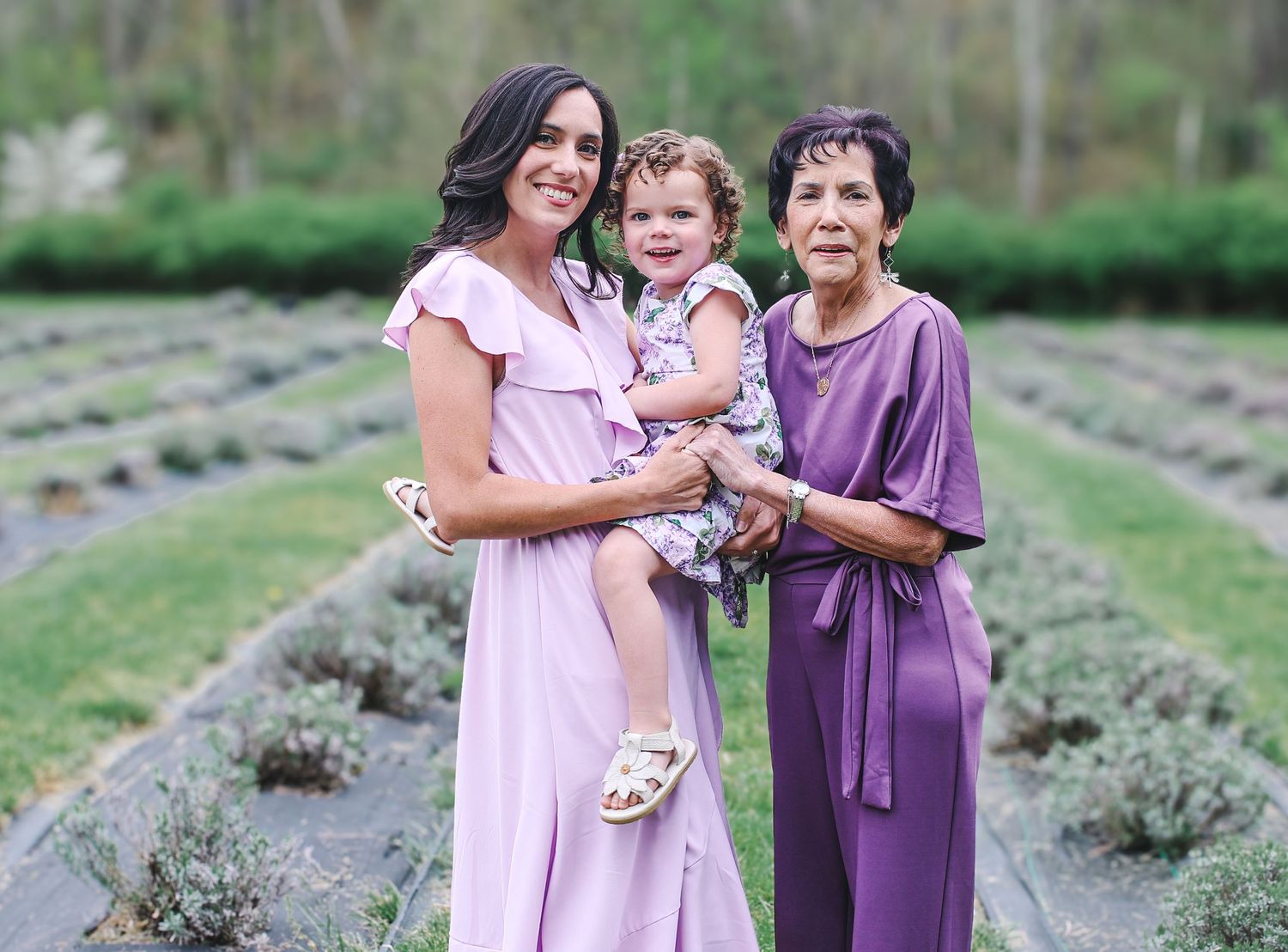 Three generations share joyful moments together in a blooming lavender field wearing coordinating purple dresses.