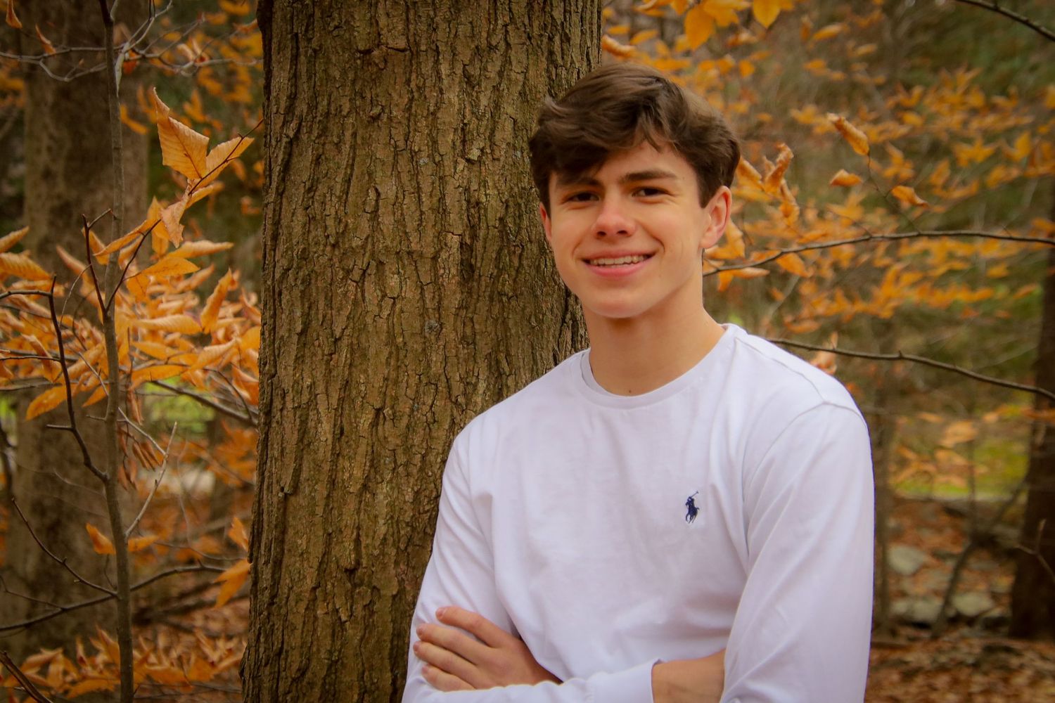 Someone in a white shirt poses against autumn foliage and tree trunks in a forest setting.